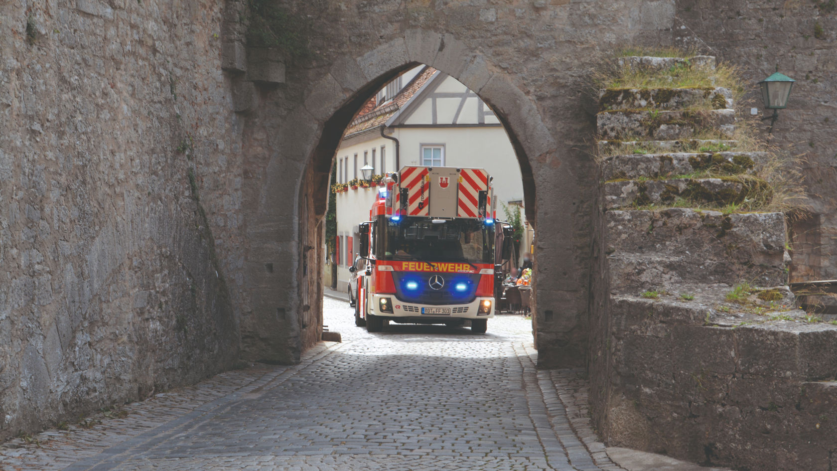 A red Econic with blue lights on driving through an old city gate on a cobbled road. The aerial rescue platform is retracted. The blue lights of the Unimog are switched on and historic buildings can be seen in the background. 