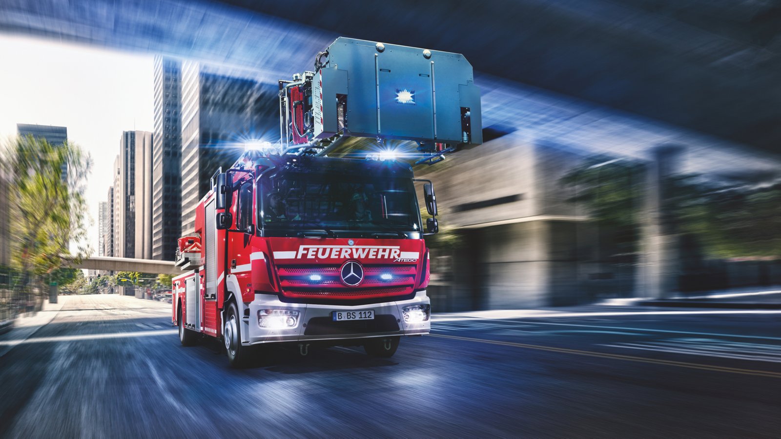 A red Atego fire-fighting vehicle drives quickly through a city with its blue lights on. The ladder of the vehicle is extended. High-rise buildings and a bridge can be seen in the background. The image conveys a sense of urgency and movement.
