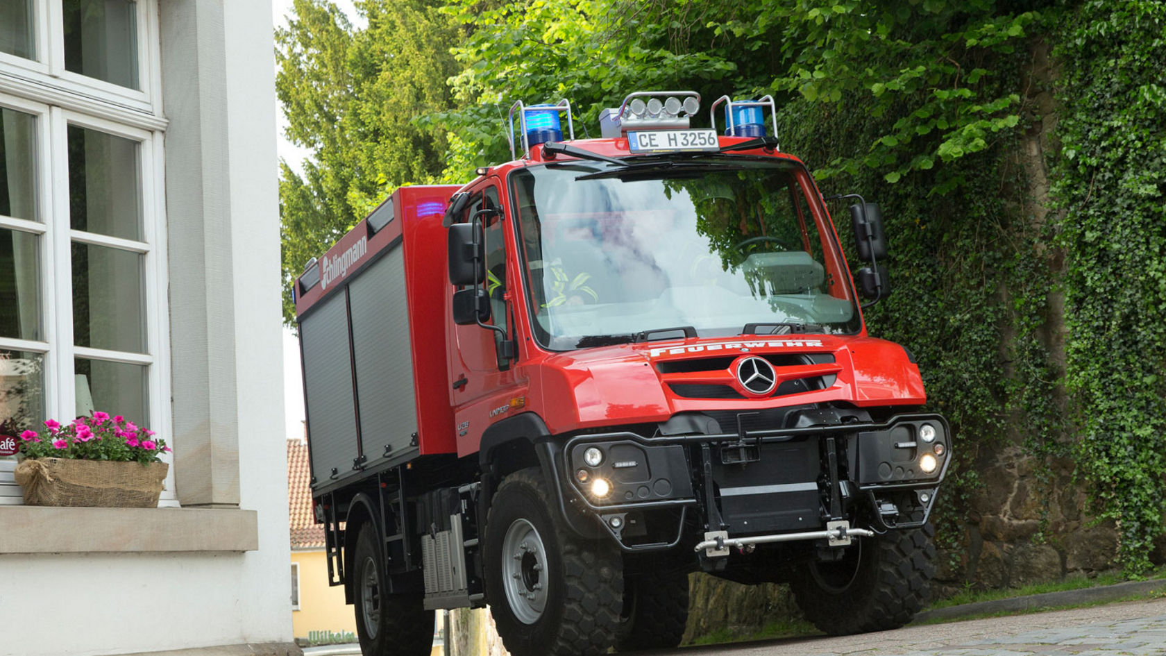 A red fire service Unimog with blue lights on driving along a narrow cobblestone road that passes a house. The vehicle is compact and manoeuvrable, ideal for use in built-up areas. “Schlingmann” lettering on the side of the Unimog indicates the bodybuilder. Two firefighters are sitting in the cab. 