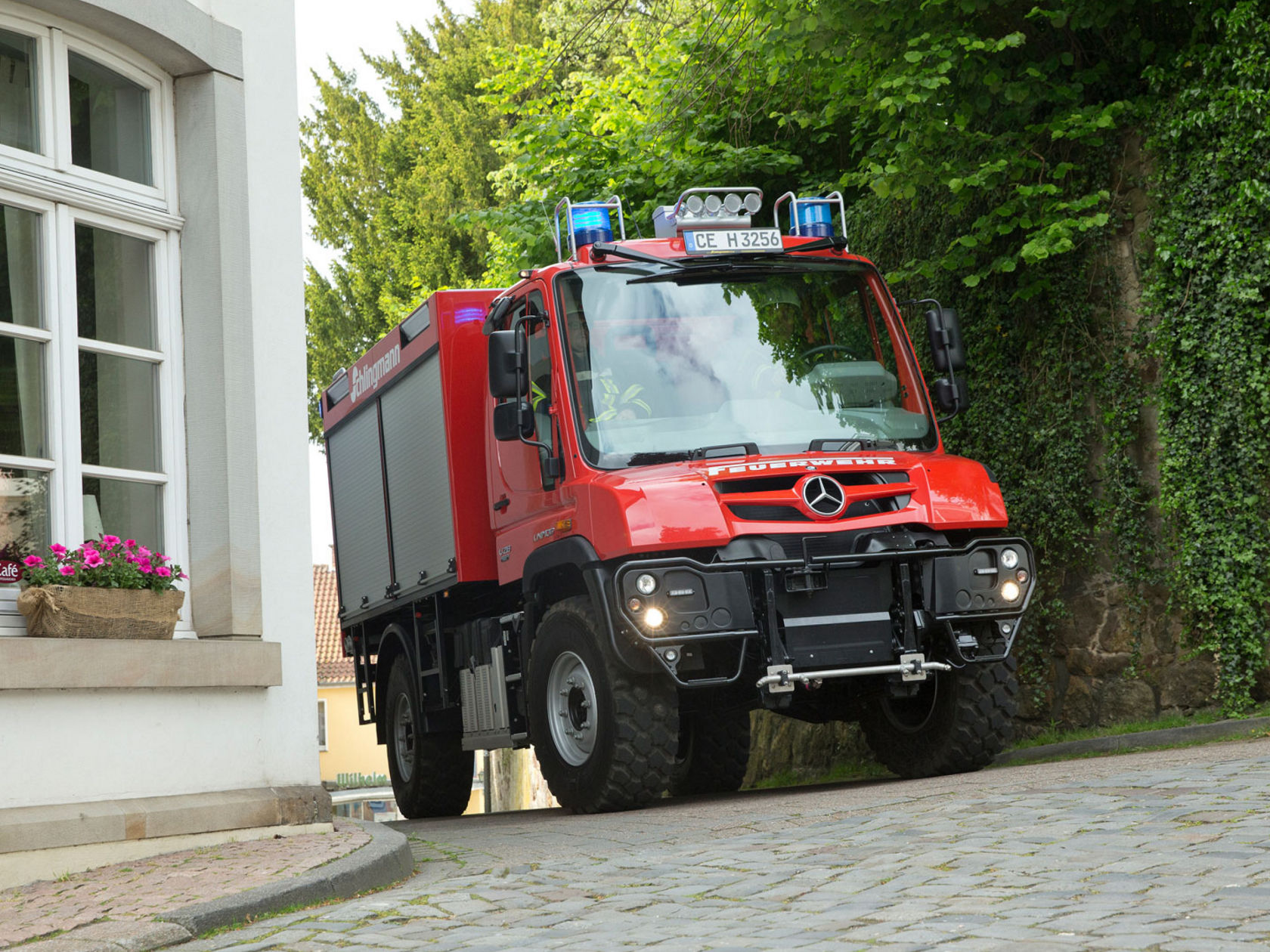 A red fire service Unimog with blue lights on driving along a narrow cobblestone road that passes a house. The vehicle is compact and manoeuvrable, ideal for use in built-up areas. “Schlingmann” lettering on the side of the Unimog indicates the bodybuilder. Two firefighters are sitting in the cab. 