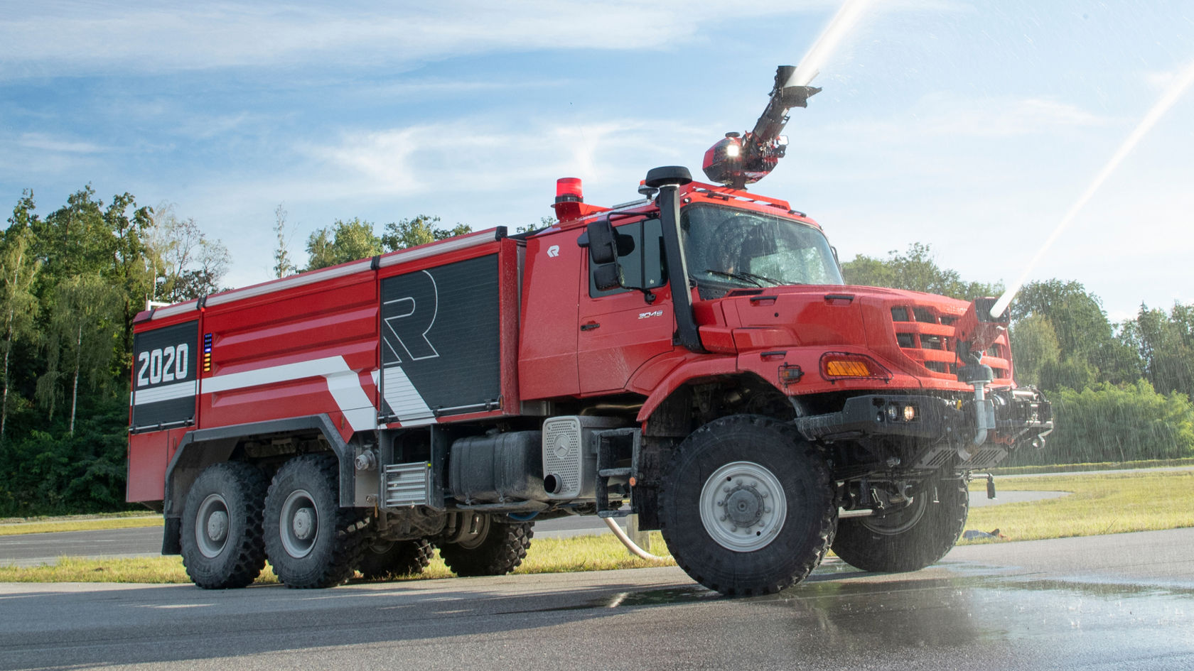 A red airfield fire-fighting vehicle sprays large quantities of extinguishing agent using its roof-mounted projector. The Zetros is equipped with large tyres and several axles, bears the lettering “2020” and a large “R”. It is located on an asphalted surface, presumably in front of an airport.