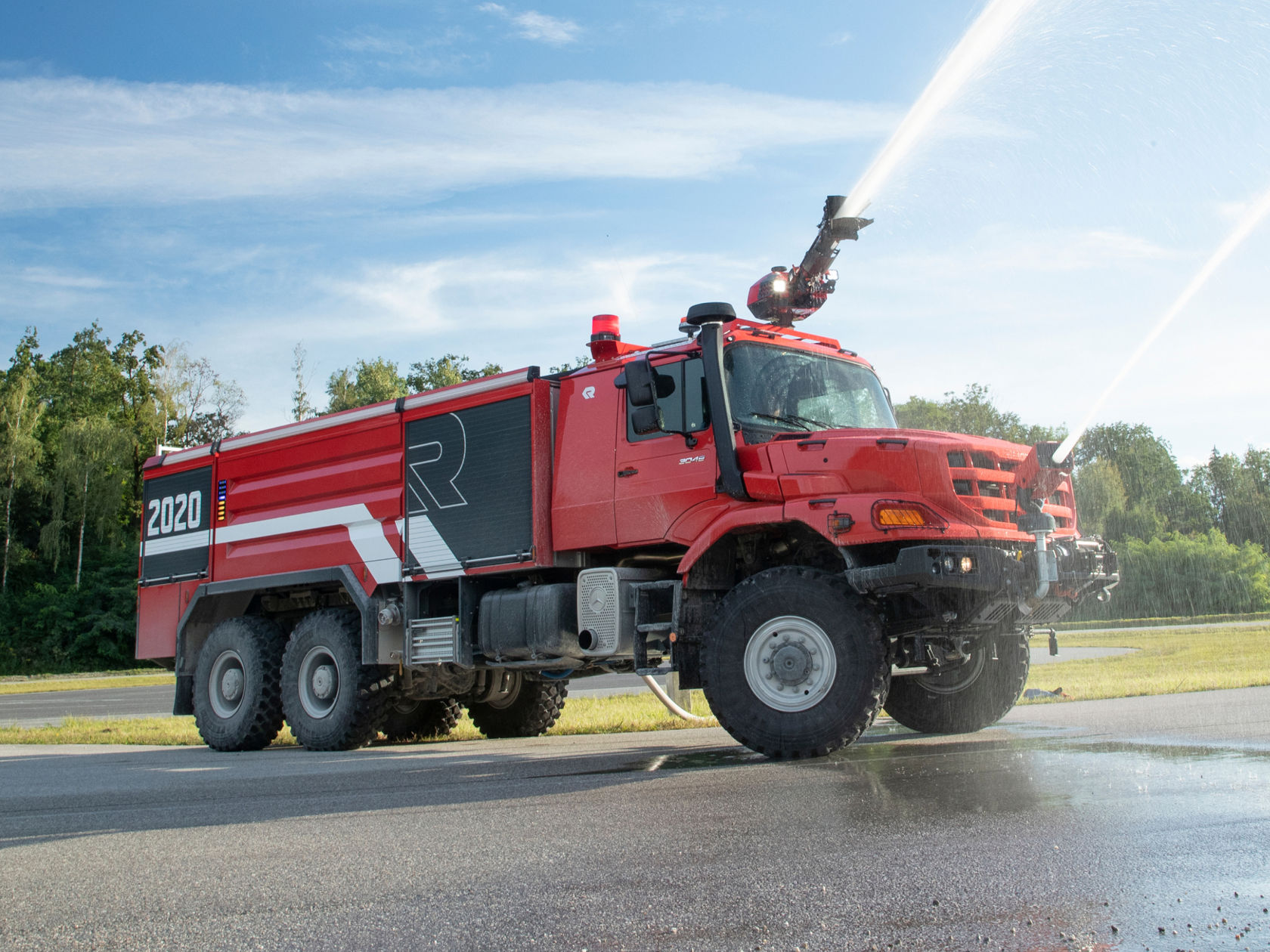 A red airfield fire-fighting vehicle sprays large quantities of extinguishing agent using its roof-mounted projector. The Zetros is equipped with large tyres and several axles, and says “2020” and a large “R”. It is located on an asphalted surface, presumably in front of an airport.