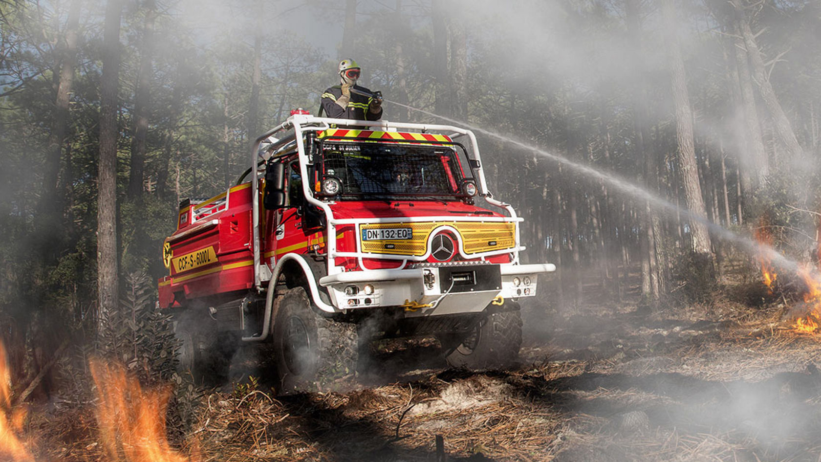 A red Unimog putting out a forest fire using its roof cannon. The forest fire-fighting vehicle in a burning forest and a firefighter in protective clothing operating the water cannon through the roof hatch. There is a protective cage in front of the windscreen.