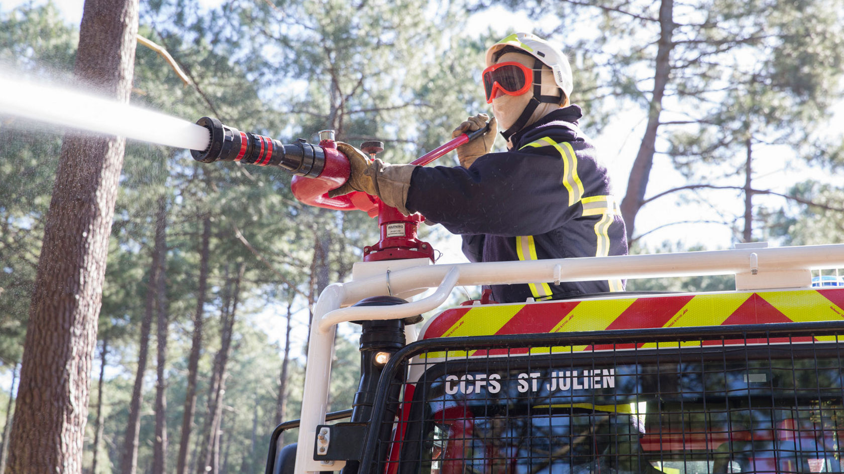 A firefighter in protective clothing including helmet, goggles and face shield operating a water canon on a fire-fighting vehicle. The jet of water shoots into the left half of the image; trees are visible in the background. The vehicle has “CCFS ST JULIEN” written on it.