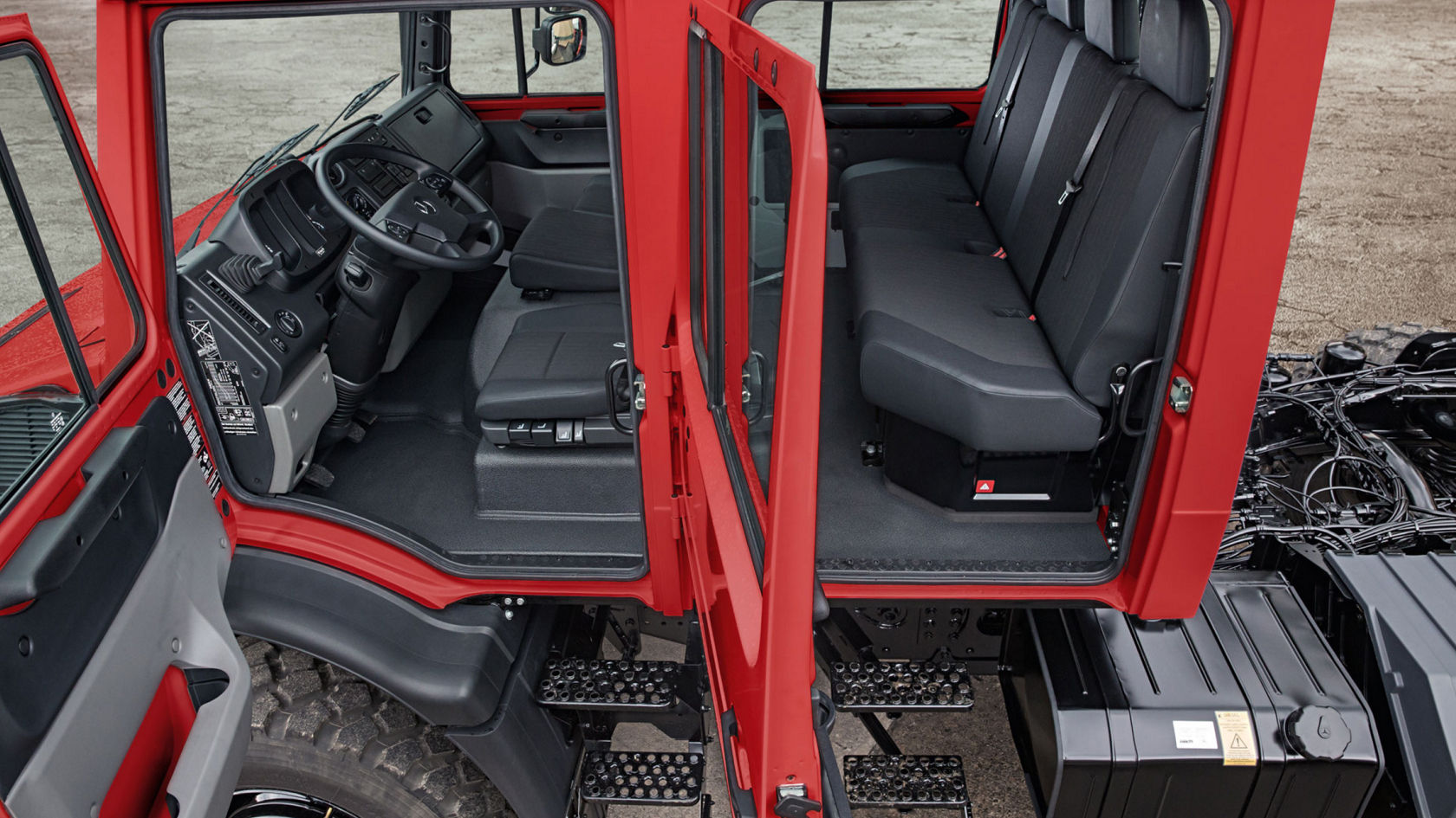 The image shows the cab of a red Unimog with open doors. This model is known for its excellent off-road capability. Inside the cab you can see the driver’s seat with steering wheel and instrument panel, behind it is a bench seat and the steps for the entrance. The interior is completely black.