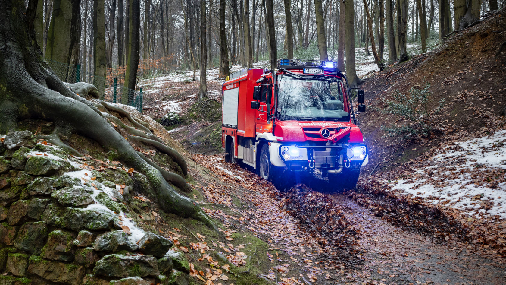 The image shows a red off-road Unimog on a muddy forest road with blue lights switched on. The ground is covered with leaves and some snow. The thick roots of a tree protrude from the slope next to the path, which is shored up by a stone wall.