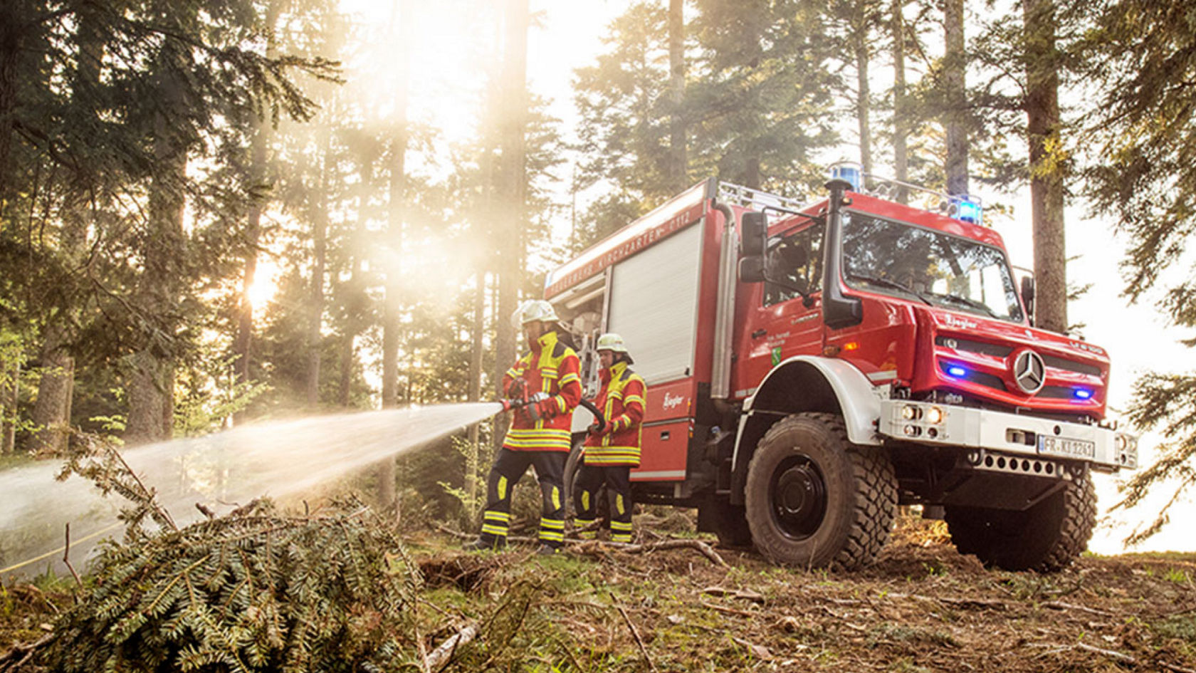 The image shows a red Unimog tanker truck in operation in the forest. Two firefighters wearing protective clothing use a hose to extinguish a small fire or embers on the ground. The Unimog standing on a forest path surrounded by trees in the sun.