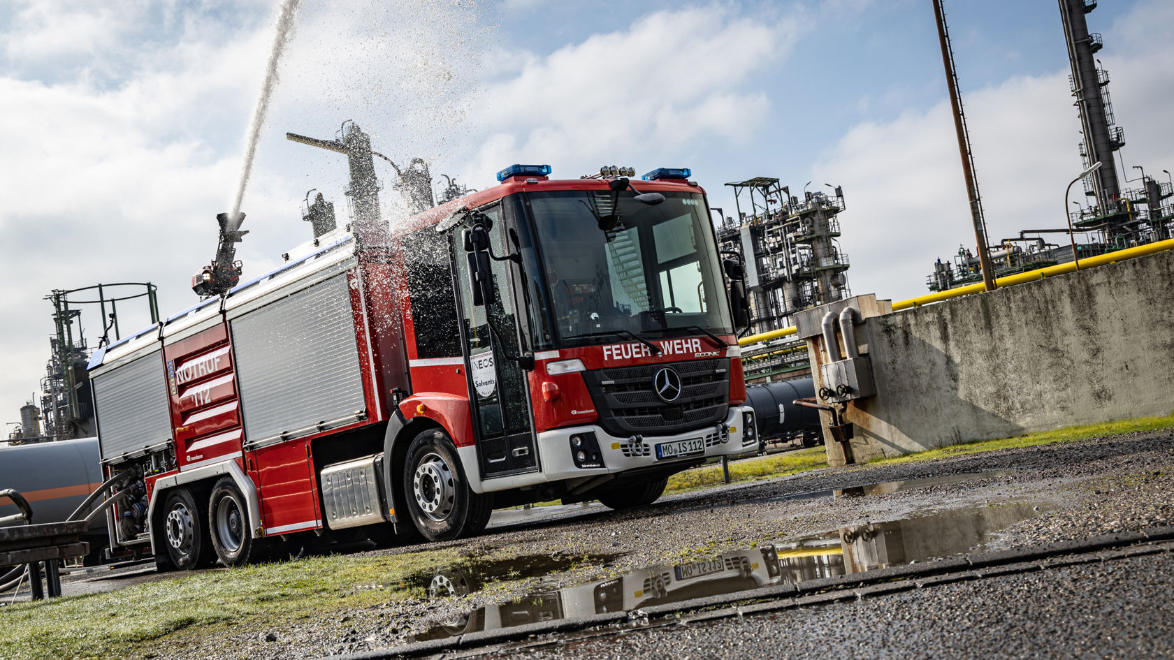 A red Econic fire truck is in operation on the premises of an industrial plant. The vehicle has an extendable water cannon on the roof firing a jet of water high into the air. The industrial plants can be seen in the background.