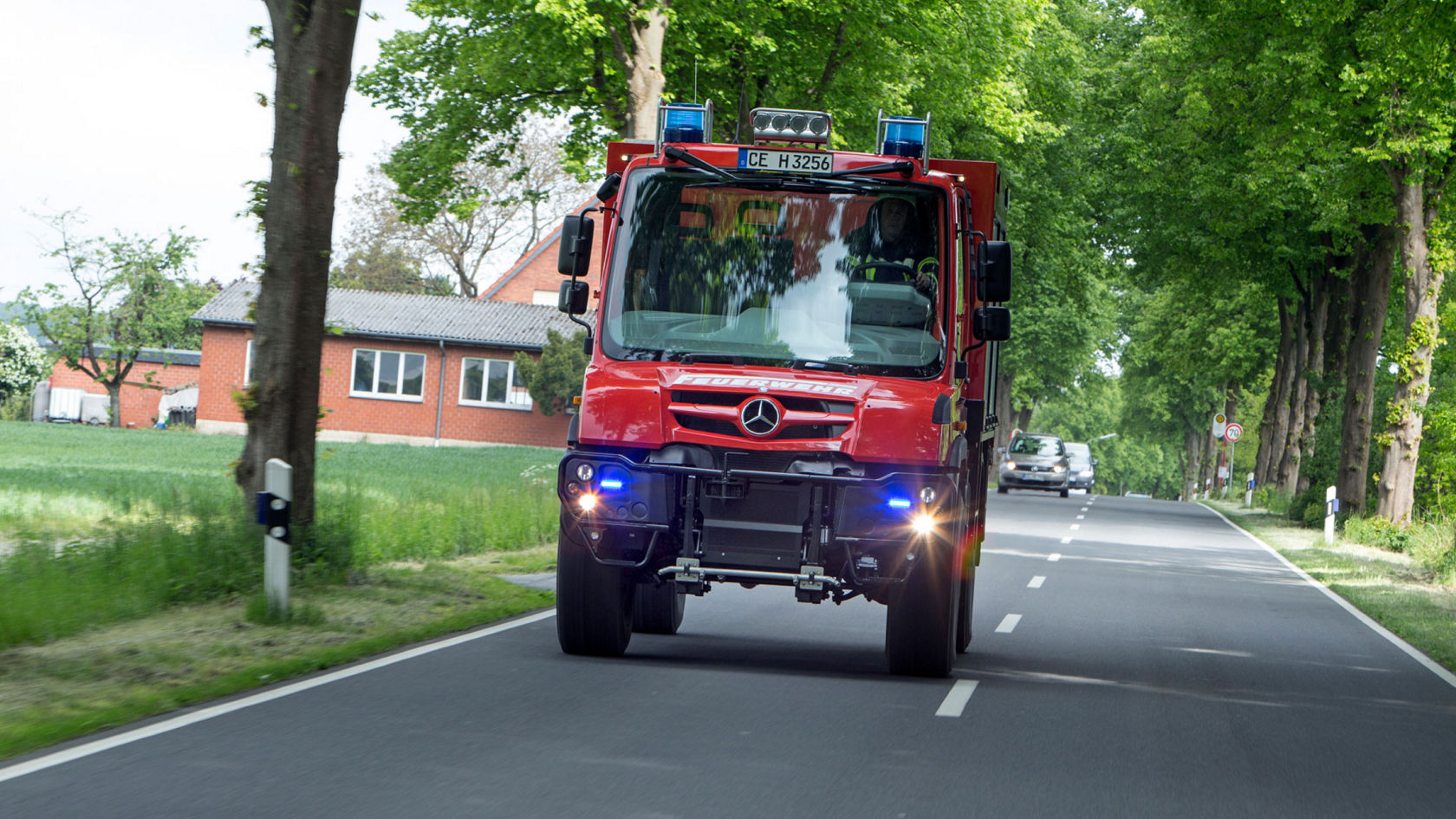 The image shows a red fire service Unimog driving on a road surrounded by trees. A red brick building and a green meadow can be seen in the background. Two cars are driving behind the Unimog some distance away.