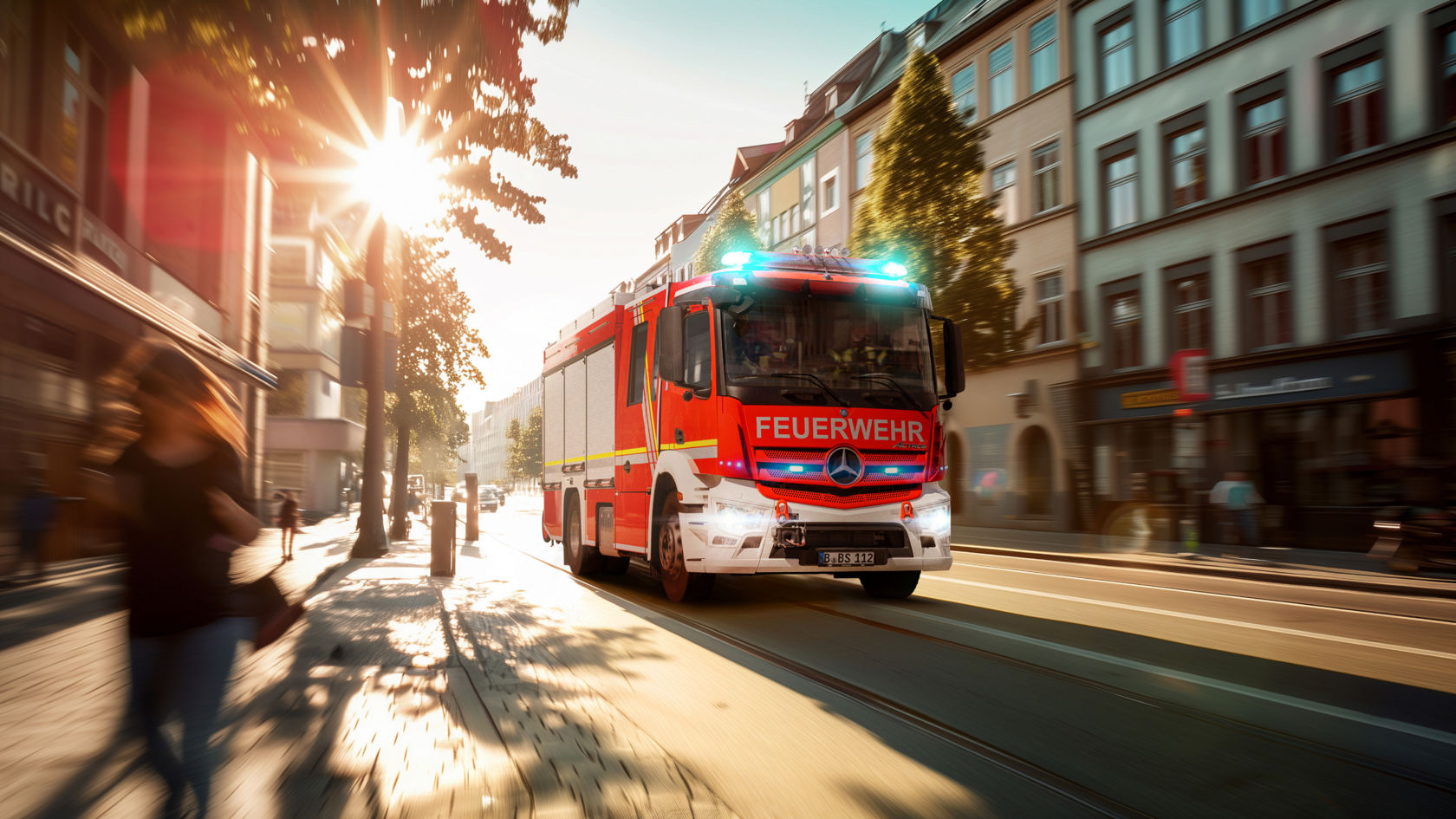 The image shows a red Actros fire-fighting vehicle driving through a city with its blue lights on. The sun shines brightly and casts long shadows on the road. Buildings and trees are visible in the background. A person is walking along the pavement.