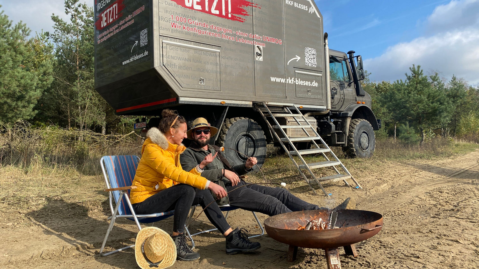 Two people are sitting at a campfire outdoors. A Unimog stands in the background.