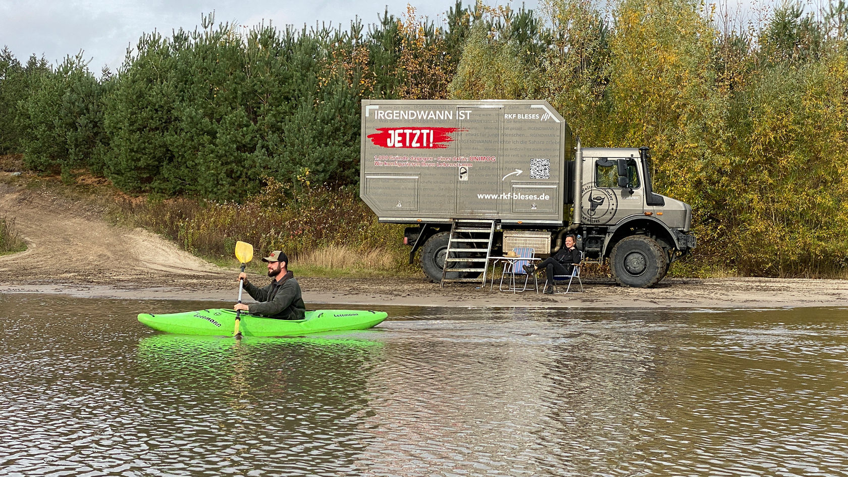 A person paddles in a green kayak on calm water, while another person sits at a table next to a large vehicle.