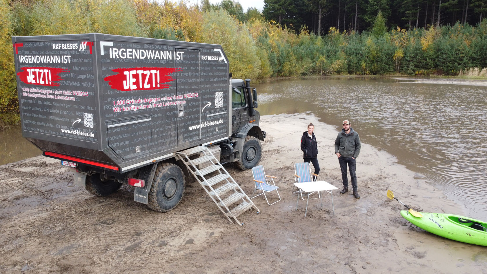 A Unimog with a camper body is standing on the shore of a lake. Two people, a man and a woman, are standing next to the vehicle.