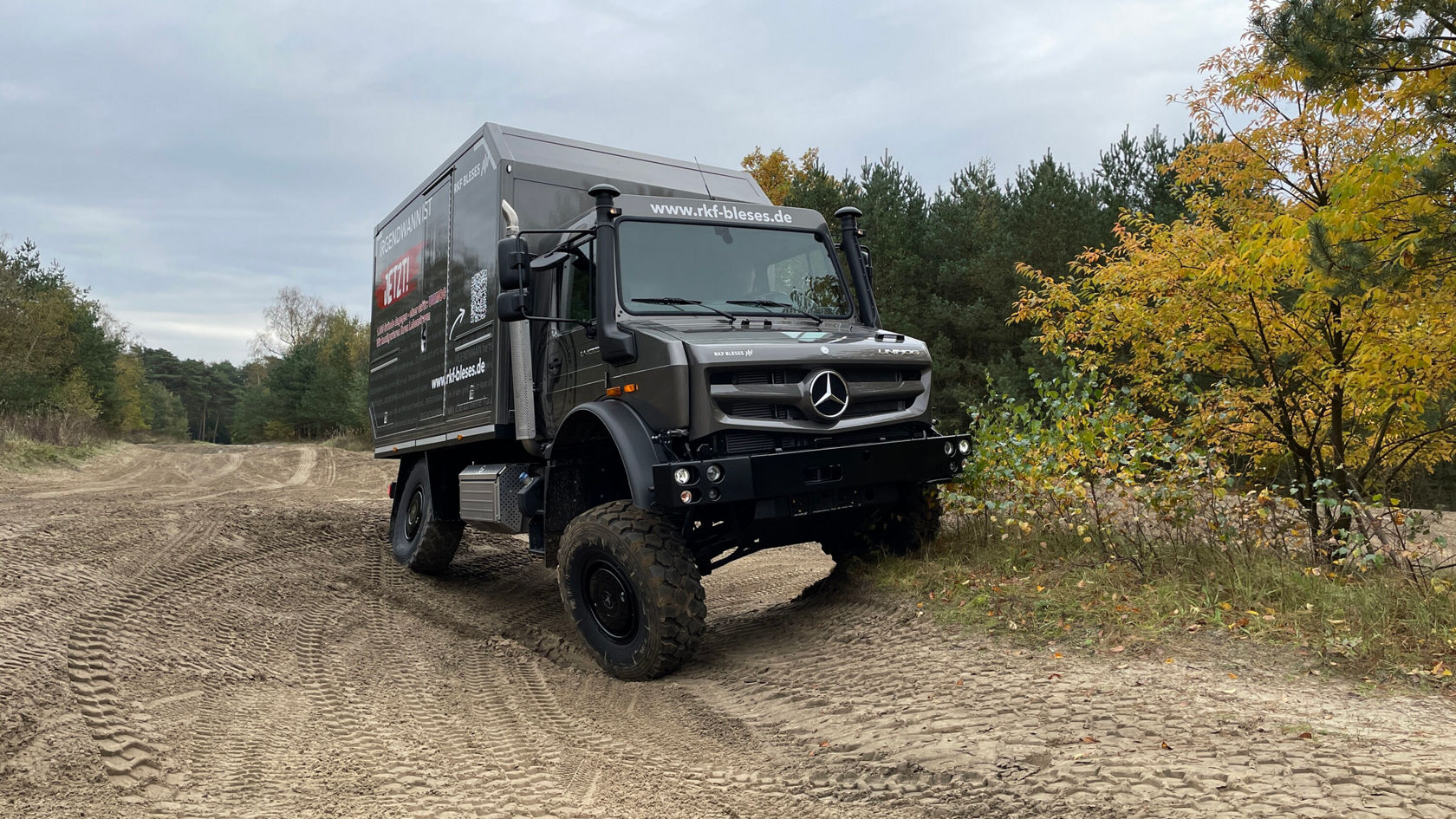 An off-road-capable Unimog from Mercedes-Benz is driving on an unpaved road surrounded by trees and shrubs.