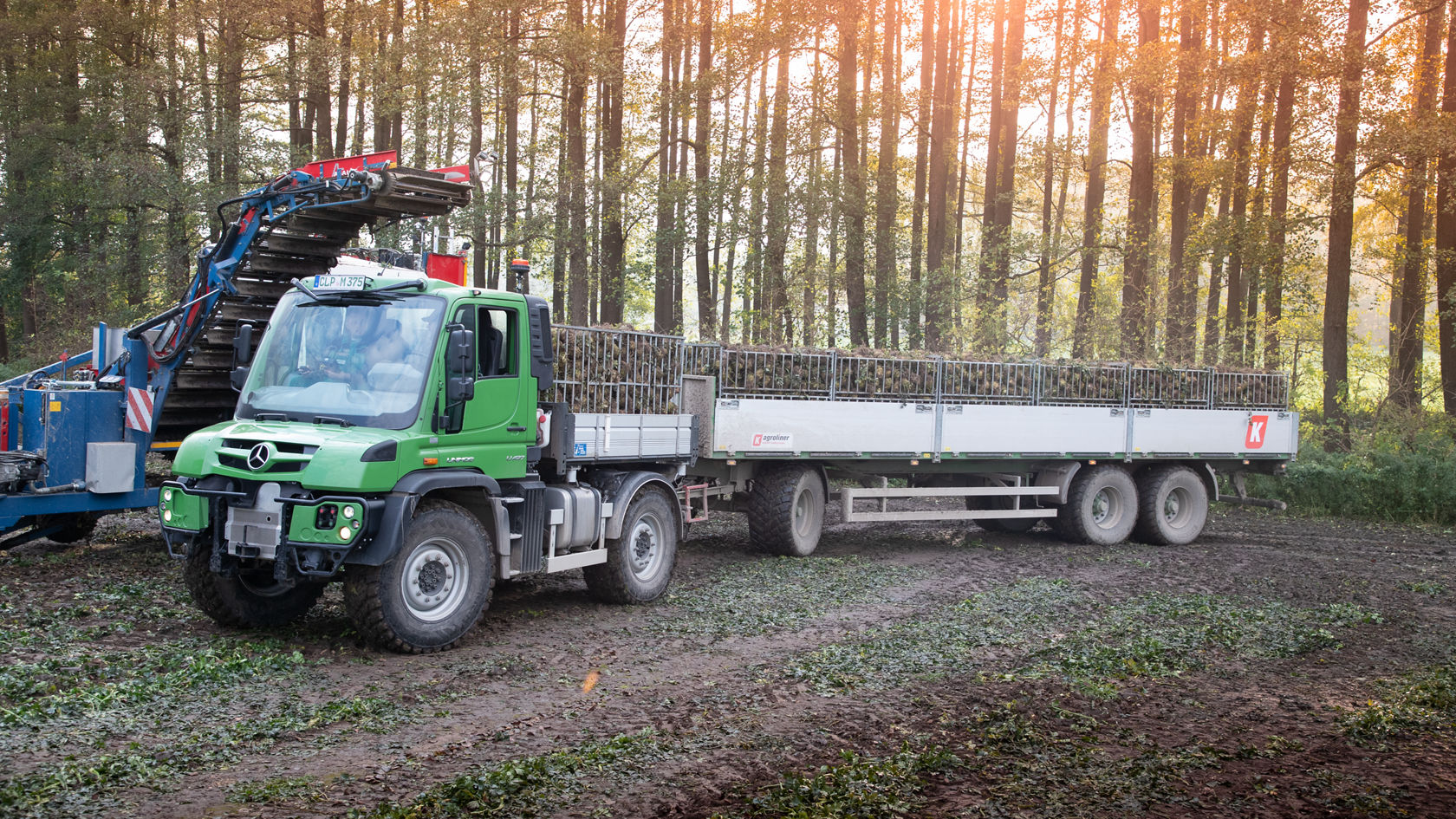 Un Unimog Mercedes-Benz vert avec une remorque se trouve dans la forêt, tandis qu'en arrière-plan une machine transporte des troncs d'arbres. La scène montre des activités forestières.