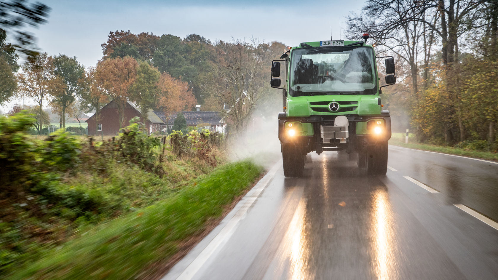 Un Unimog Mercedes-Benz vert roule sur une route de campagne humide, entouré d'arbres aux couleurs automnales et avec une propriété rurale en arrière-plan.