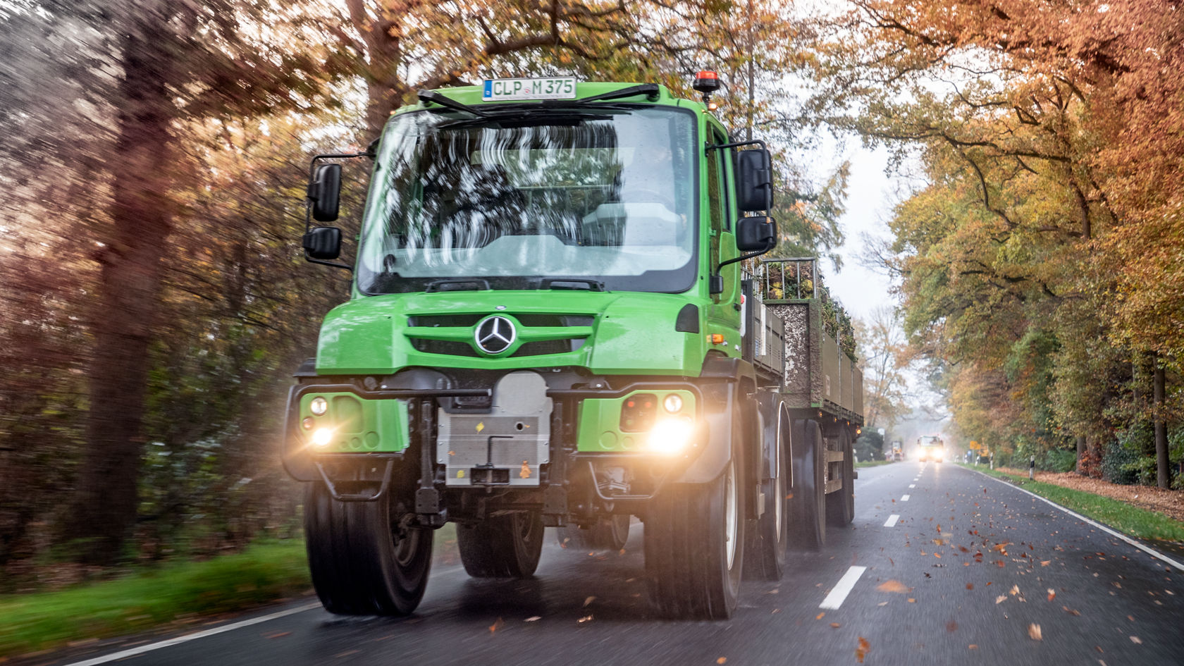 Un camion Mercedes‑Benz vert roule sur une route de campagne, entouré d'arbres aux couleurs automnales. Les phares sont allumés et il est équipé d'un attelage. Un camion Mercedes‑Benz vert roule sur une route de campagne, entouré d'arbres aux couleurs automnales. Les phares sont allumés et il est équipé d'un attelage.