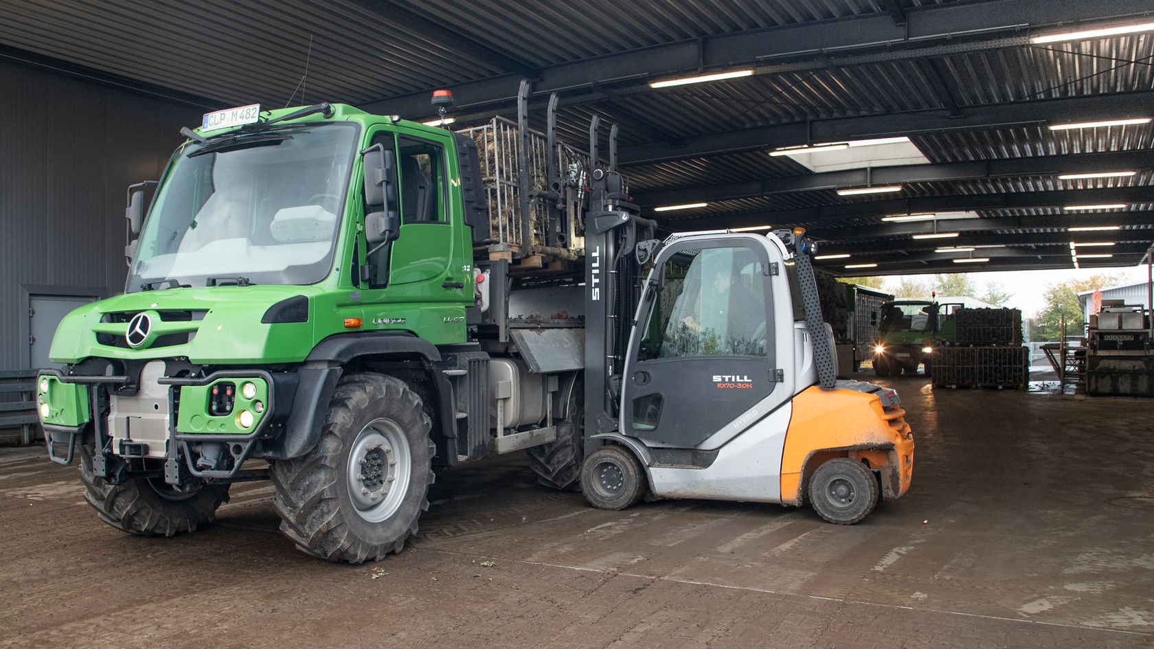 Un Unimog Mercedes‑Benz vert se trouve à côté d'un chariot élévateur dans un entrepôt. Le chariot élévateur soulève une palette de marchandises.