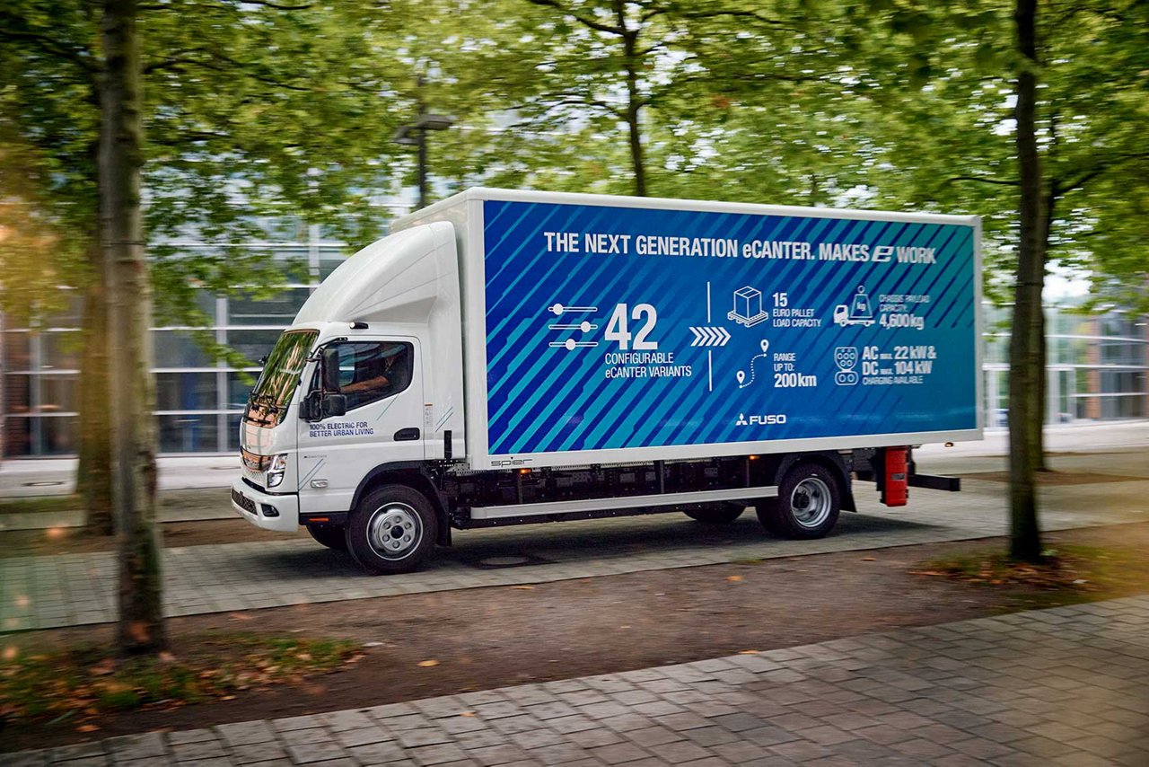 weißer Fuso Ecanter Lkw fährt auf einer Straße zwsichen grünen Bäumen durch. Er hat eine Aufschrift auf blauem Hintergrund.
