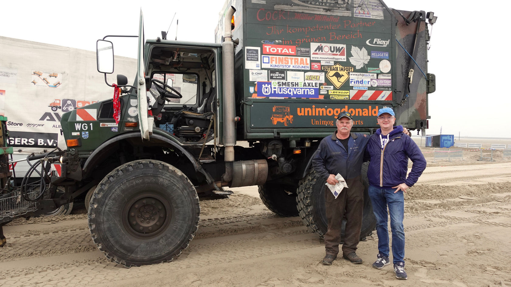 Zwei Männer stehen neben einem Mercedes-Benz Unimog, der mit verschiedenen Aufklebern versehen ist, auf einem sandigen Untergrund.