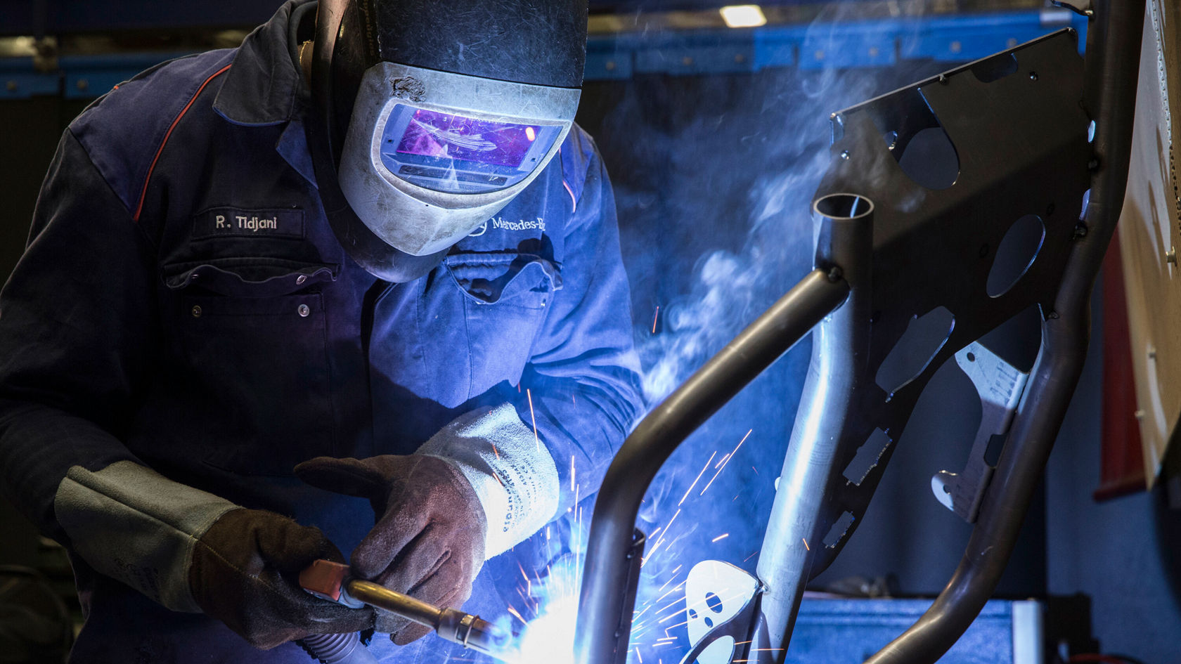 Ein Mechaniker schweißt einen Metallrahmen mit einer Schutzschweißmaske, während Funken sprühen. Der Fokus liegt auf dem Schweißgerät und dem glühenden Metall.
