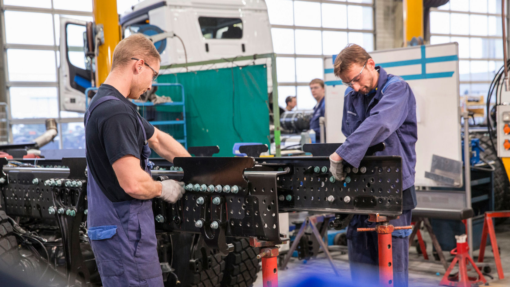 Zwei Mechaniker arbeiten in einer Werkstatt an einem Fahrzeugrahmen, während sie Teile mit Schrauben und Werkzeugen montieren. Im Hintergrund ist ein größerer Lkw sichtbar.