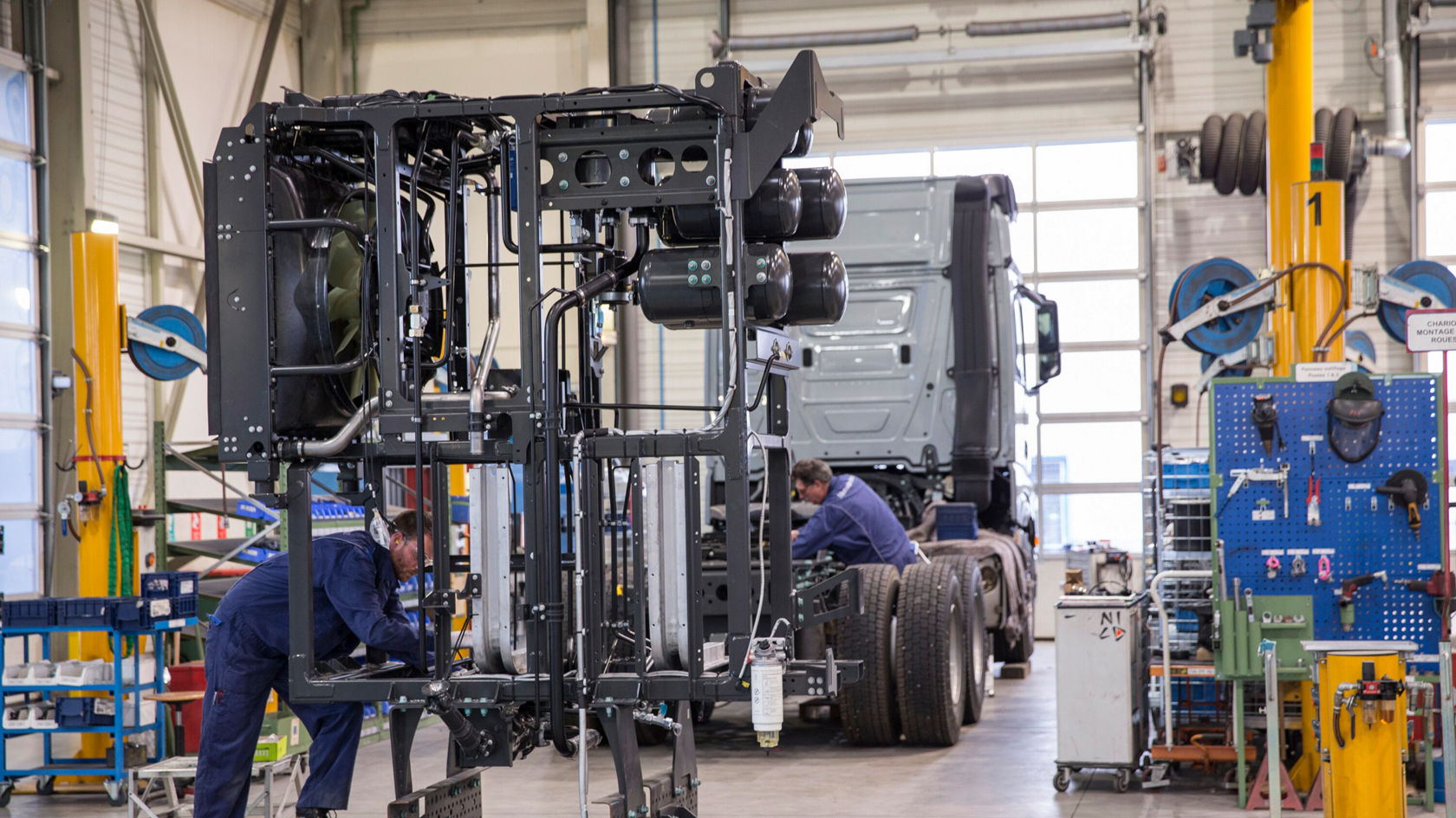 Zwei Mechaniker arbeiten an einem Mercedes-Benz Lkw-Chassis in einer Werkstatt, während ein weiterer Lkw im Hintergrund sichtbar ist.
