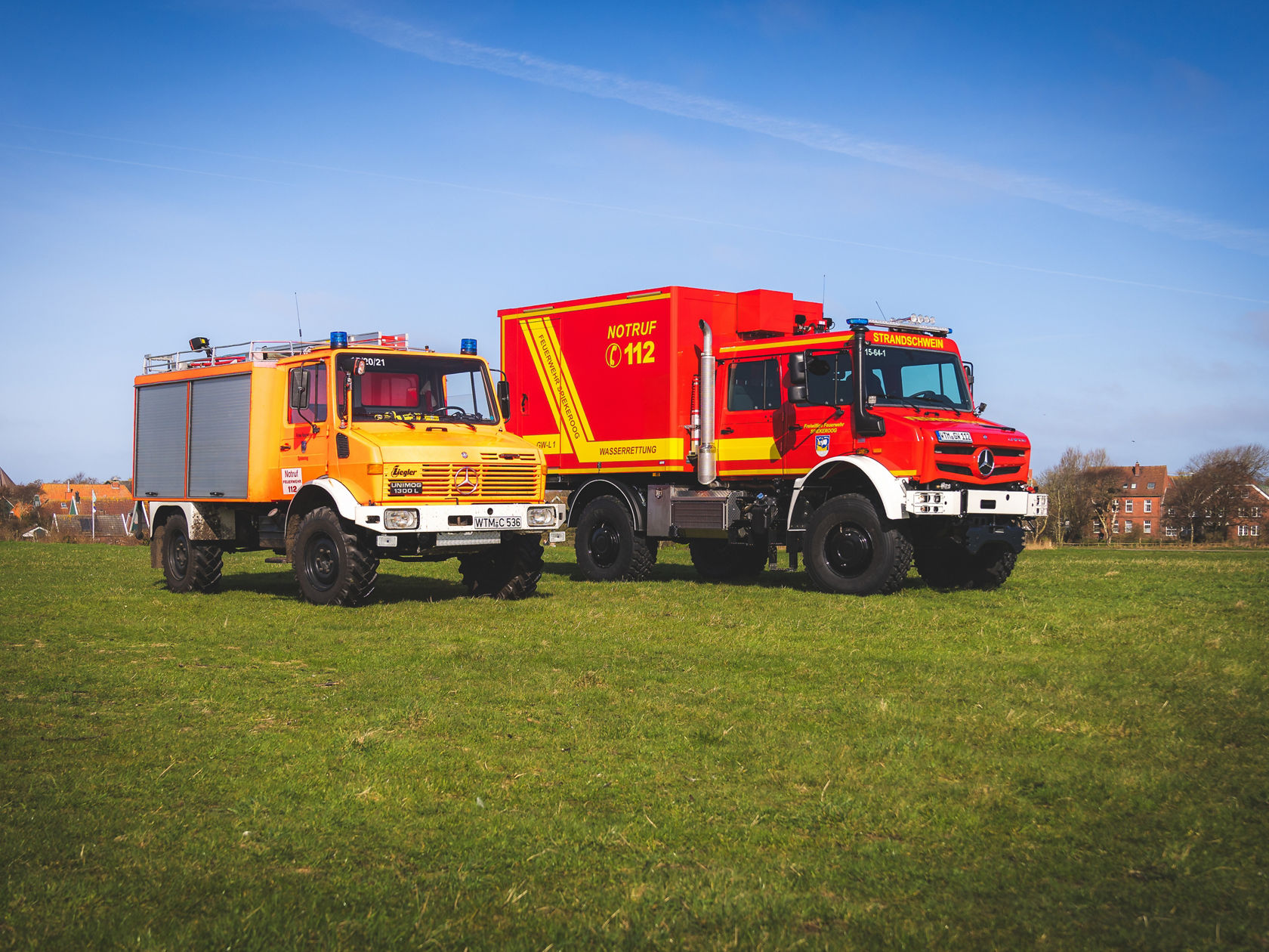 Ein Unimog 1300 L und ein Unimog U 5023, beide in Feuerwehr-Ausführung, stehen auf einer Wiese