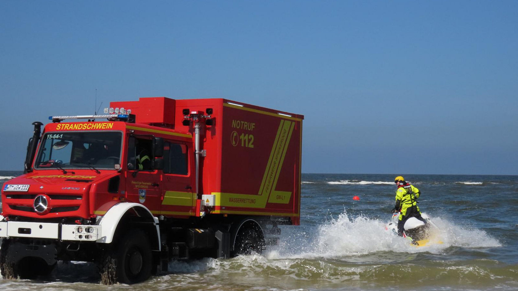 Ein Feuerwehr Unimog steht im Wasser. Eine Einsatzkraft sitzt auf einem motorisierten Wasserrettungsfahrzeug.