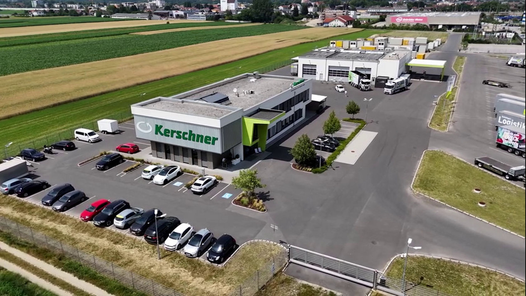 Kerschners Firmengebäude ist aus der Vogelperspektive zu sehen. Auf dem Gelände stehen mehrere Fahrzeuge geparkt, umgeben von Rasenflächen und Bäumen. Im Hintergrund erstrecken sich Felder.