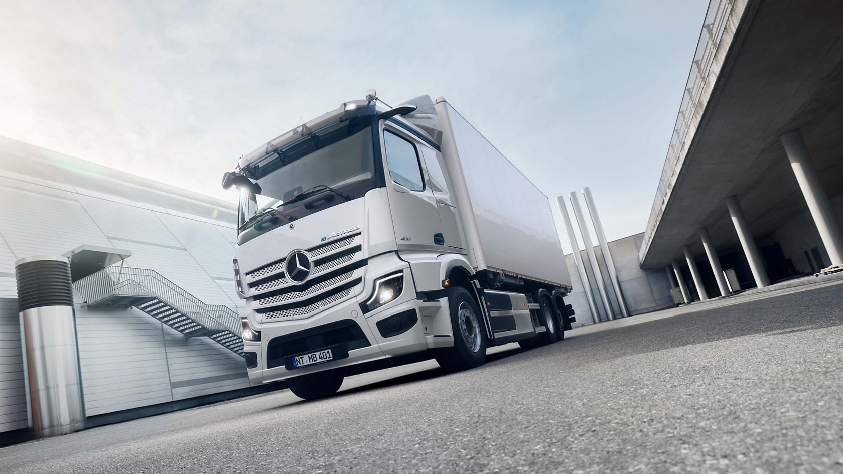 A white eActros 400 from Mercedes-Benz Trucks stands on tarmac in front of a modern building with an outside staircase, with the sun shining brightly from the top left. On the right side of the picture is an elevated concrete structure with columns indicating an underpass or a covered area.