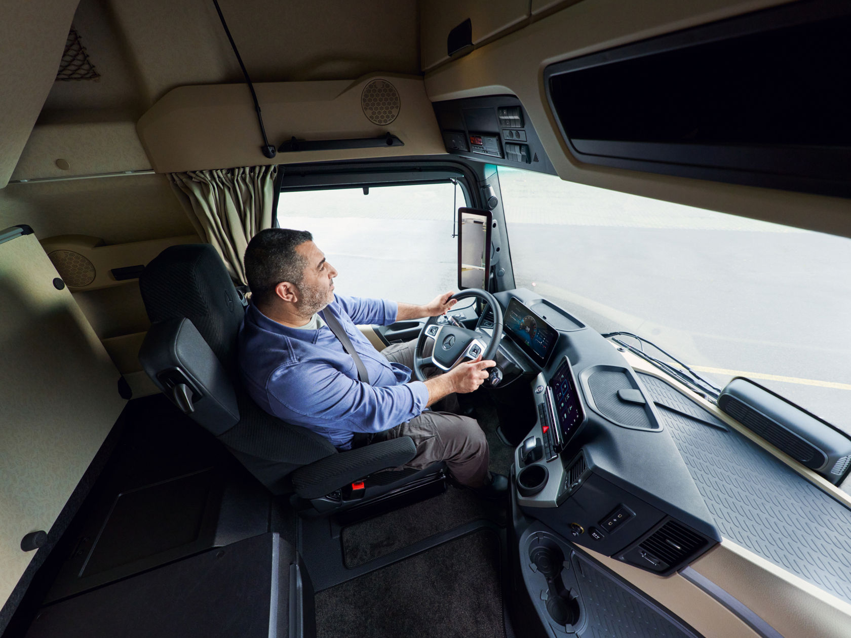 Een man met baard en blauw shirt zit op de bestuurdersstoel van een Actros L en houdt het stuurwiel vast. Hij kijkt naar rechts, terwijl de weg voor hem door de voorruit zichtbaar is.