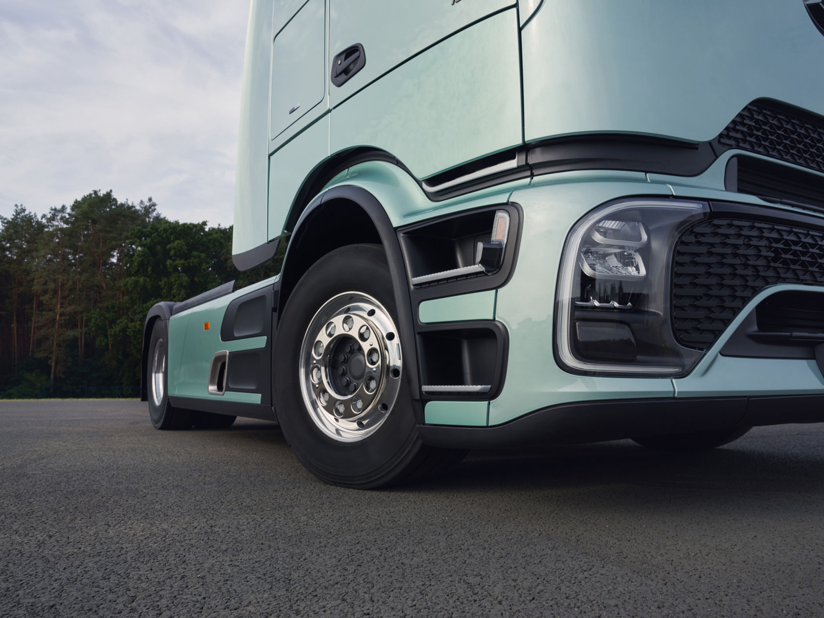 A large, mint-green Actros L stands on an asphalt surface, with the focus on the front end and the front wheel. Trees and a cloudy sky can be seen in the background.
