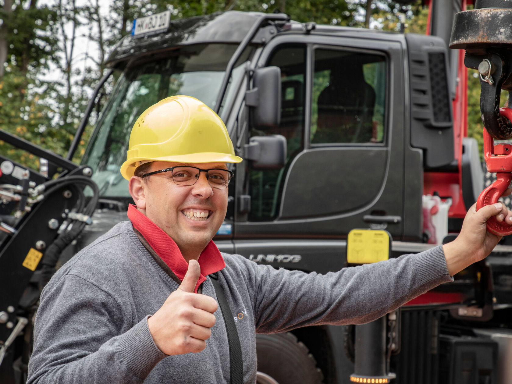 A person with a yellow safety helmet and glasses stands in front of a construction vehicle. He is giving a thumbs up.
