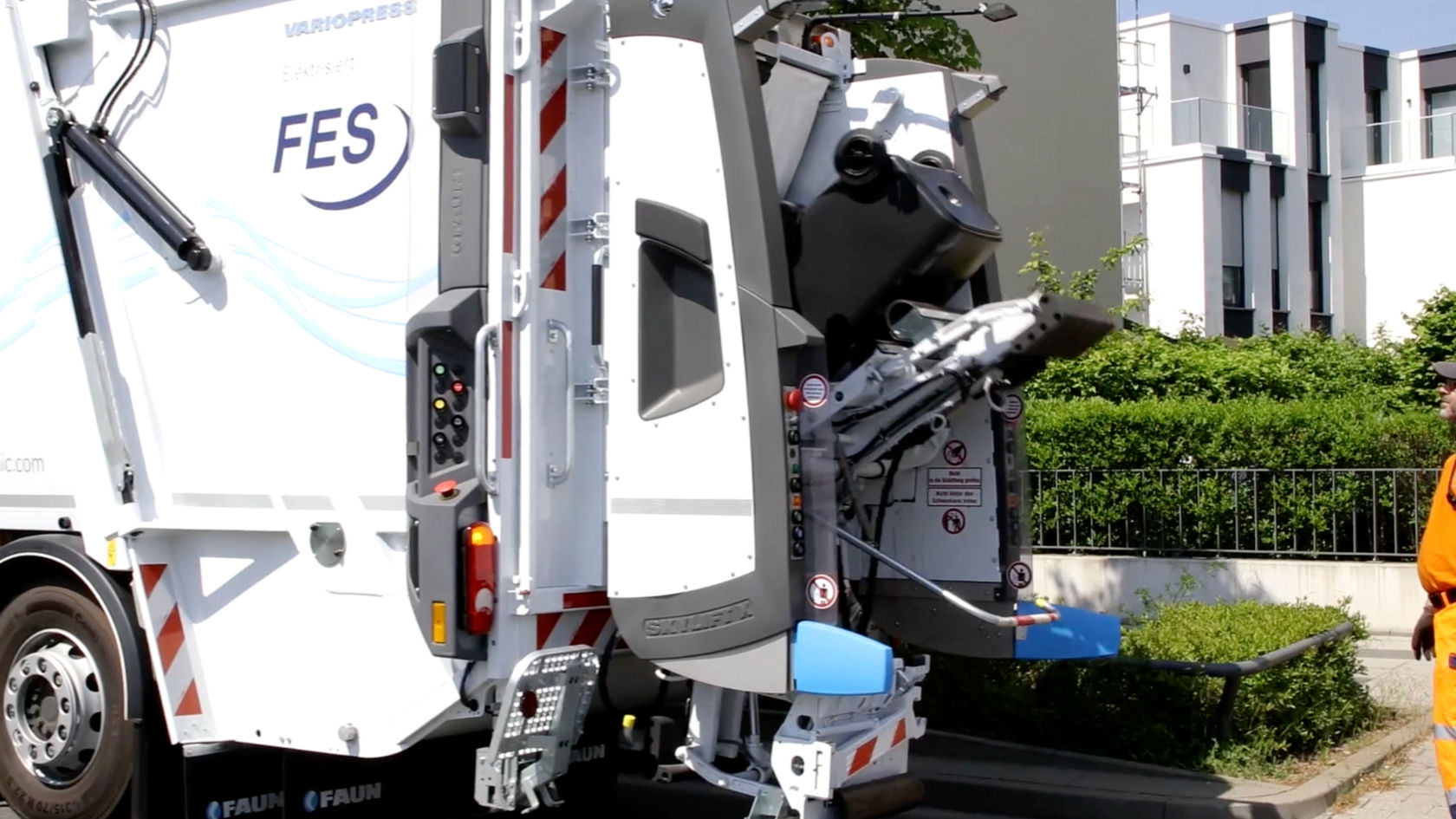 The image shows the rear of an Econic. This is a refuse collection vehicle with “FES” lettering. The truck’s lifting mechanism is extended and in motion. An employee in orange work clothes stands next to the rear of the vehicle. In the background is a modern residential building.