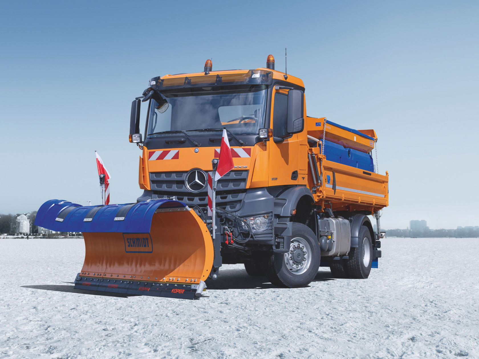 Ein orangefarbener Arocs mit Schneepflug steht auf einer verschneiten Fläche. Der Pflug ist blau und trägt den Namen "Schmidt". Der Himmel ist hellblau.