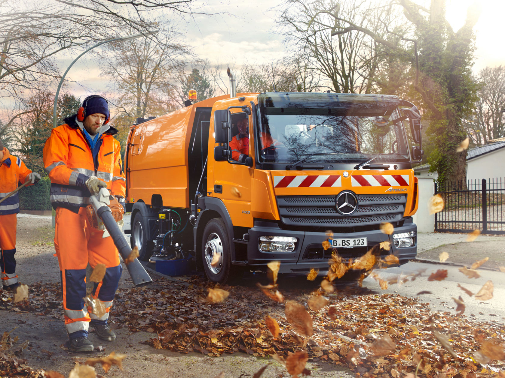 Ein orangefarbener Atego steht auf einer Straße voller Herbstlaub. Ein Arbeiter in orangefarbener Arbeitskleidung benutzt einen Laubbläser.