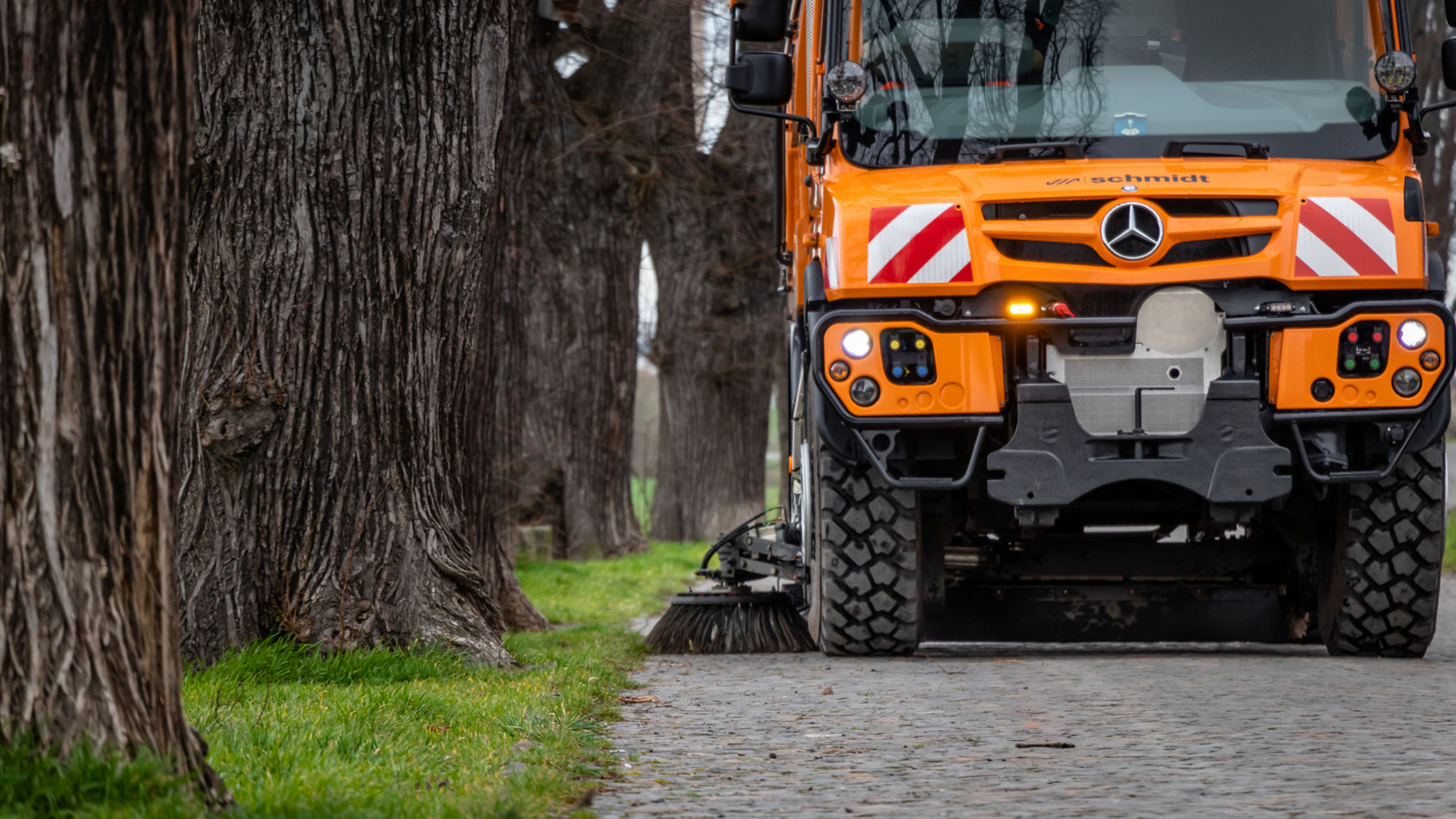 The image shows a close-up of an orange Unimog with a road sweeper on a cobbled road. The rotating brush on the side of the machine is in operation, sweeping the road. Large tree trunks and a green strip of lawn can be seen in the foreground.