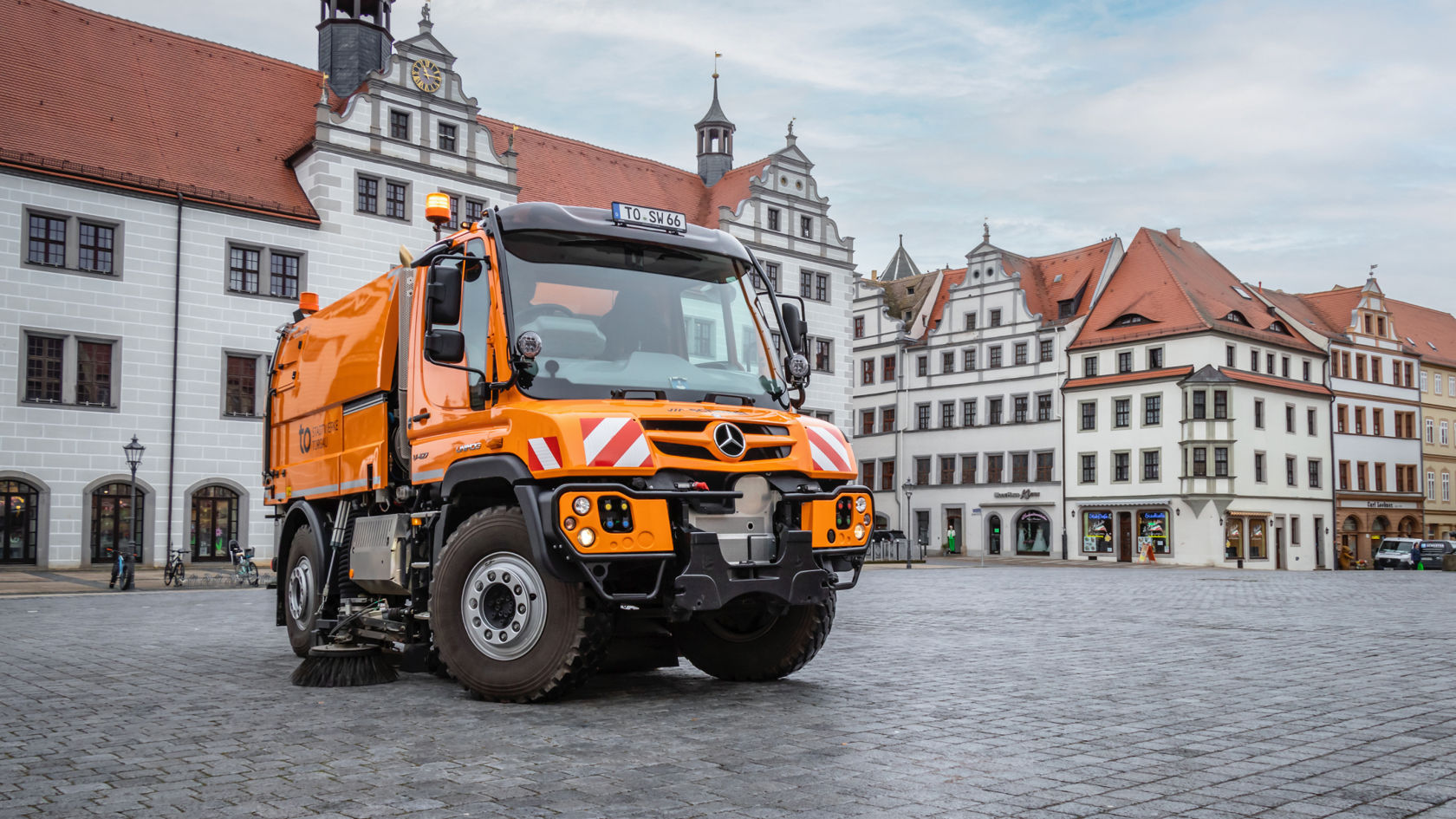 An orange Unimog with a road sweeper parked in a cobbled square. In the background are historic buildings with white façades and red roofs. The square seems empty, with just the Unimog standing there. 