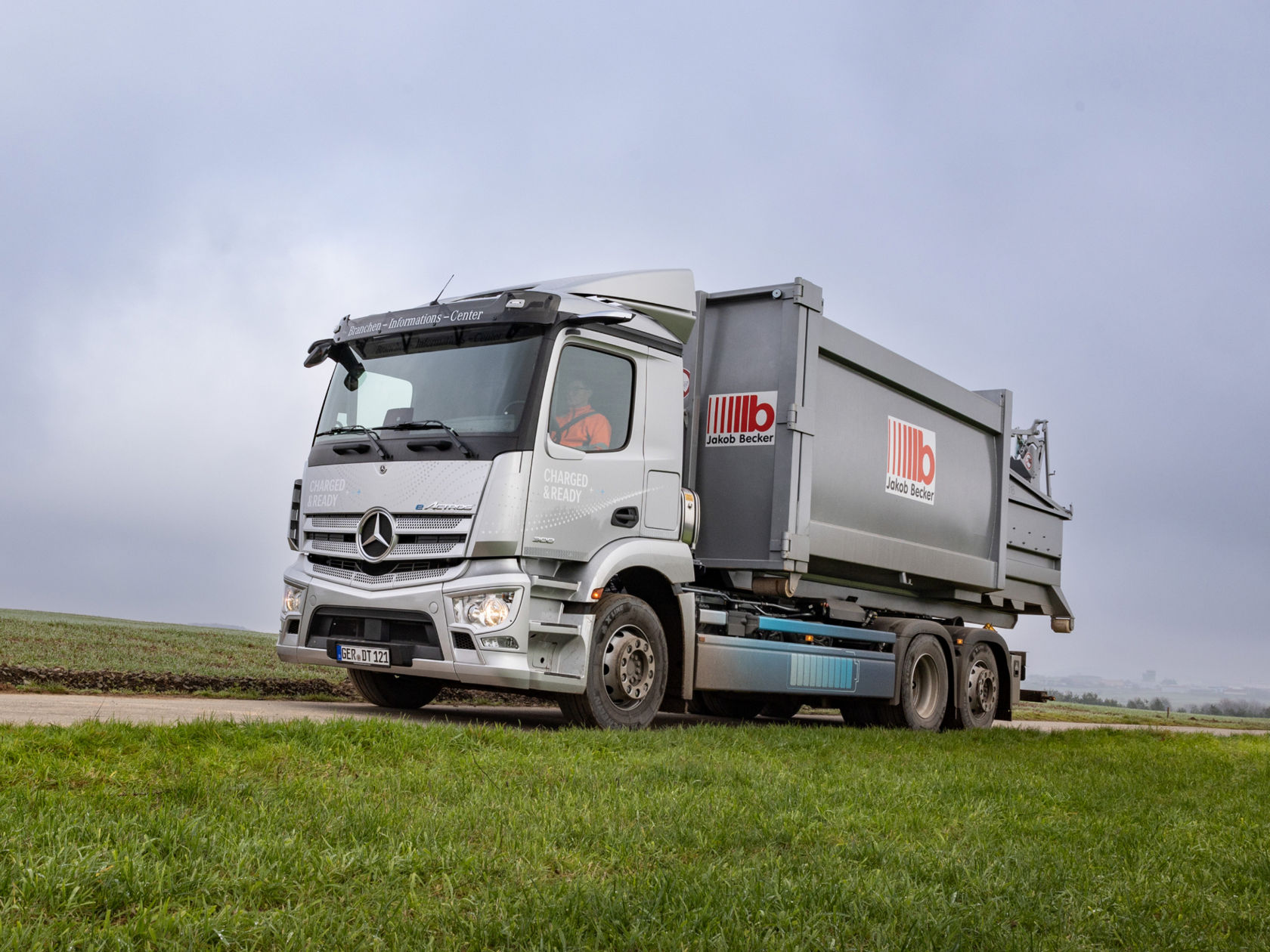 Ein silberner eActros mit "Jakob Becker" Aufschrift steht auf einer Straße neben einer grünen Wiese. Der LKW trägt den Schriftzug "Charged & Ready". Der Himmel ist bewölkt.