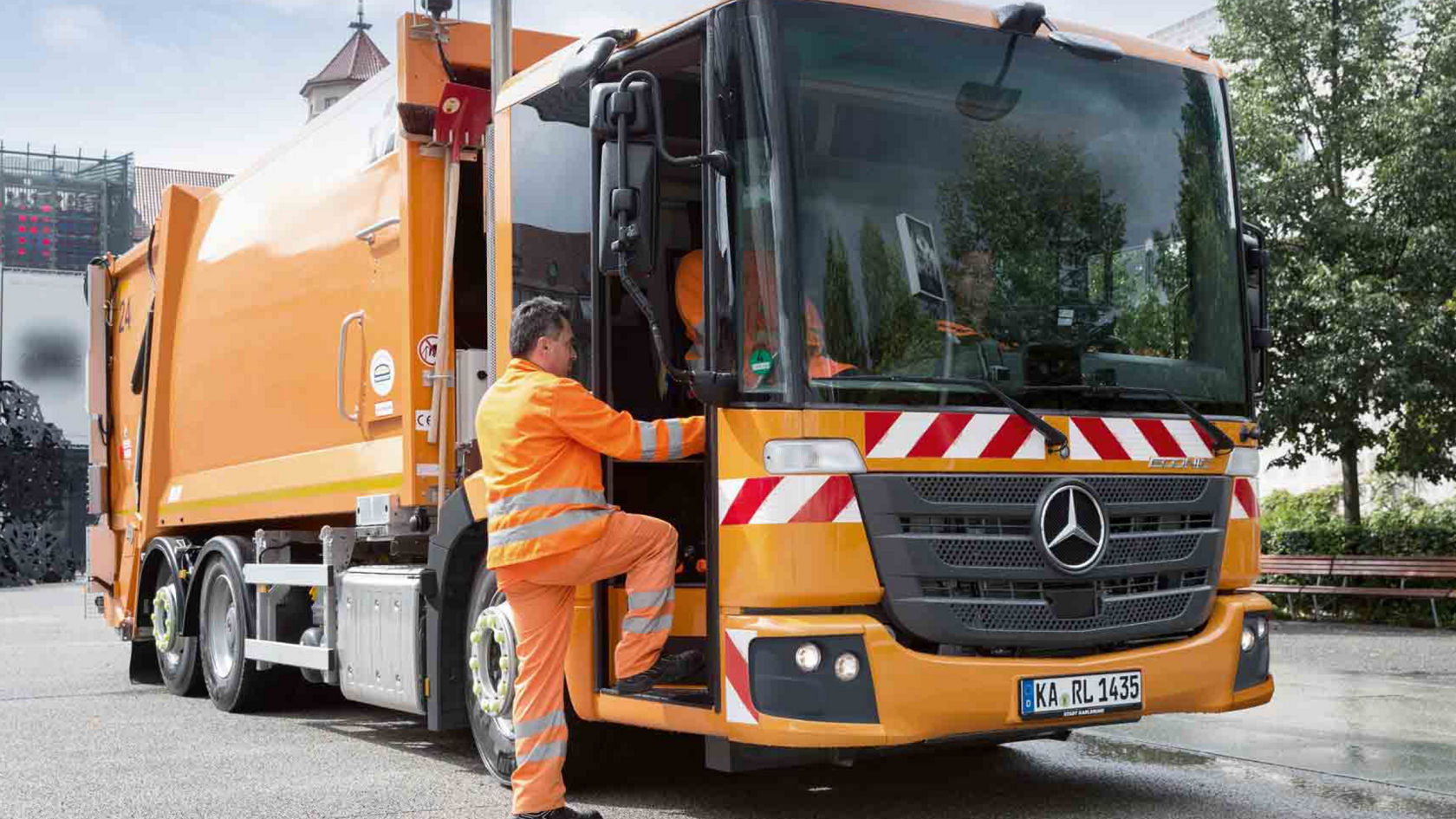 An orange Econic refuse collection vehicle is parked on a road. A waste collection worker in orange high-visibility clothing is climbing into the cab. A building and trees are visible in the background.