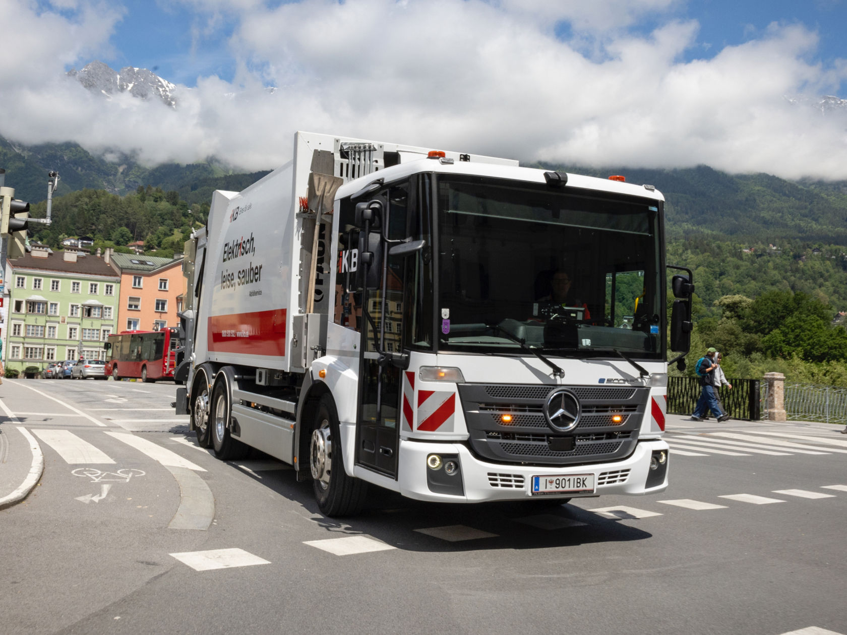 Ein weißer eEconic Müllwagen fährt auf einer städtischen Straße. Der LKW ist mit "Elektrisch, leise, sauber" beschriftet. Im Hintergrund sind Gebäude, ein Bus und Berge zu sehen. Ein Fußgänger überquert die Straße. Das Bild ist hell und klar.