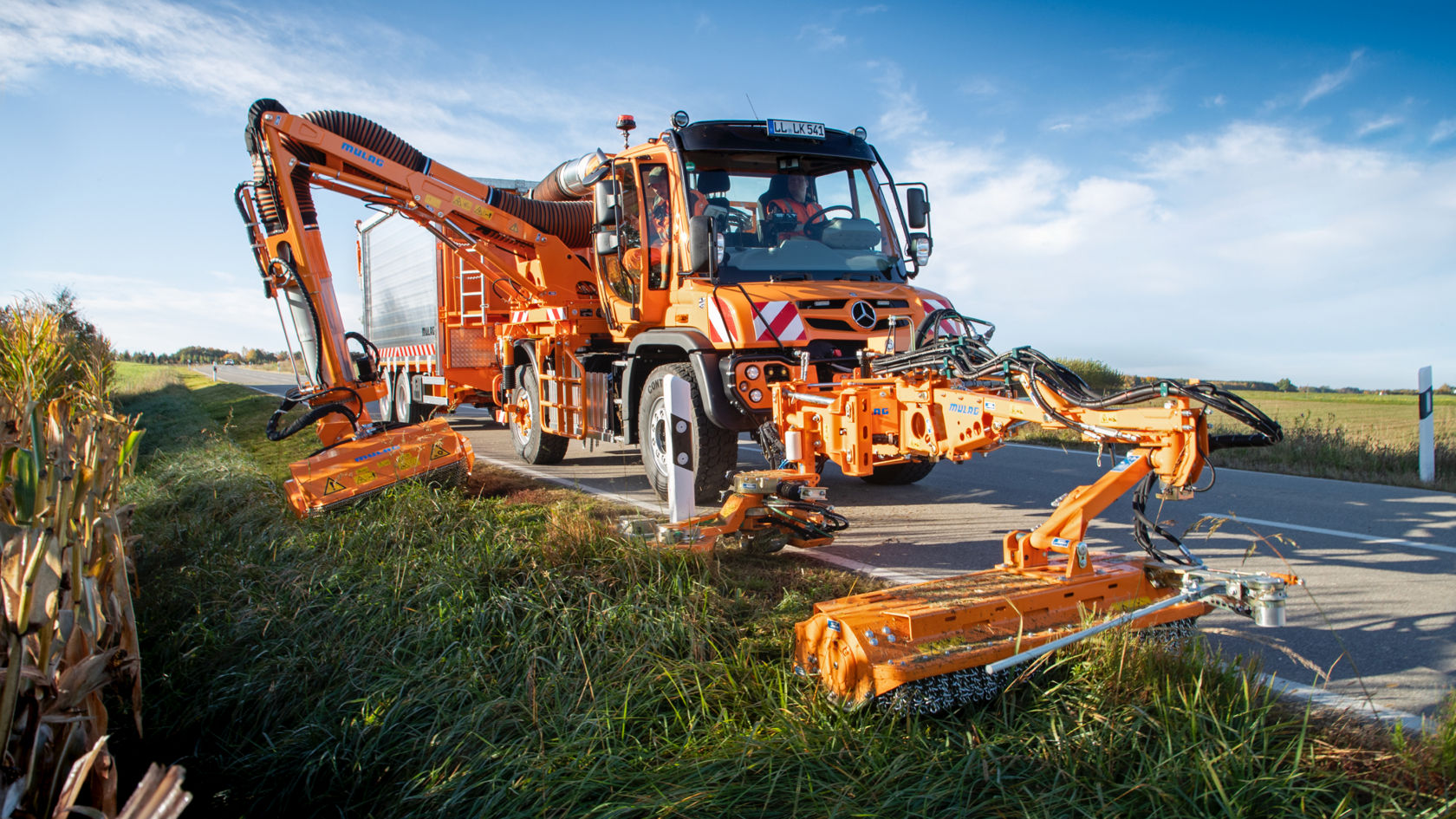 An orange Unimog with two side-extended mower decks working on the roadside. The Unimog, with a boom-like body behind the cab, is driving on an asphalt road. Tall grass and corn plants can be seen next to the road. 