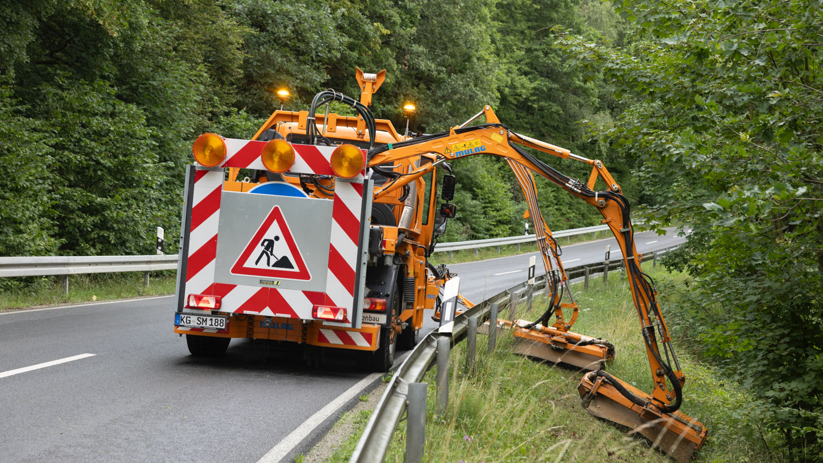 An orange Unimog with an extendable mower arm mowing by the roadside. The vehicle has a red and white warning sign and a construction site sign at the rear. The mower arm is leaning over the crash barrier and mowing the vegetated side of the slope next to the road. A forested landscape can be seen in the background.