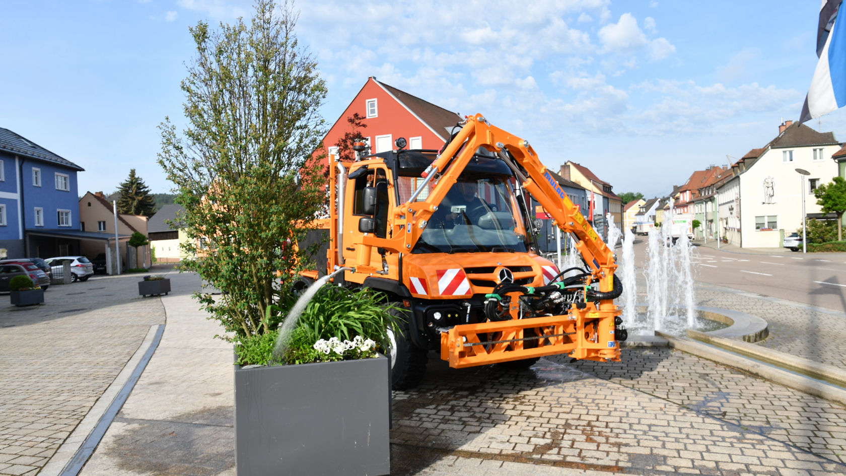 An orange Unimog with a crane arm mounted at the front of the truck watering a planter on a cobbled square. The Unimog has a snowplough linkage at the front, but it is not in use. In the background is a fountain, houses and parked cars. 