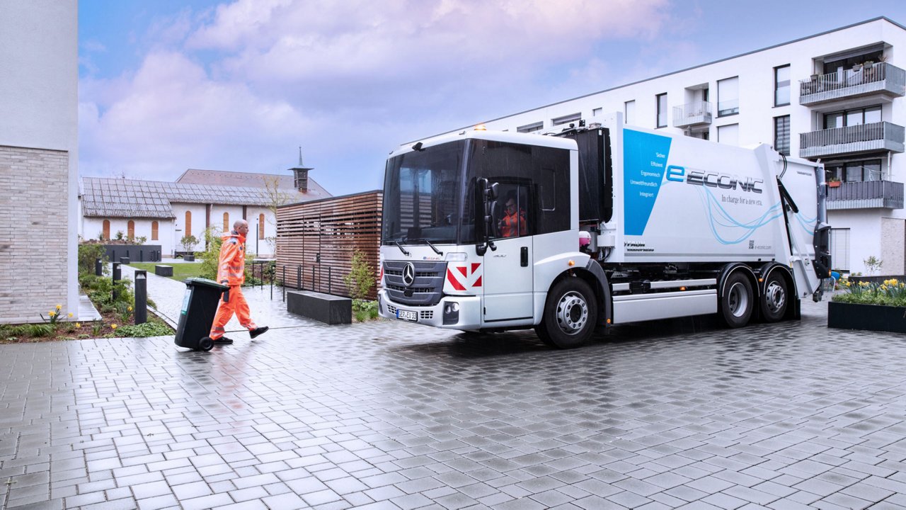 A white Econic refuse truck is standing on a cobbled courtyard. An employee in orange work clothes is pulling a bin towards the vehicle. In the background are modern residential buildings and a building with a wooden façade. The sky is cloudy and the ground is damp. 