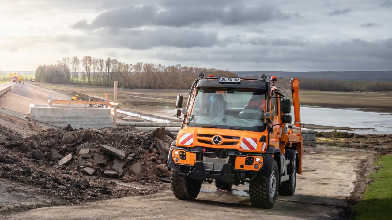 An orange Unimog driving alongside a body of water on a construction site. A heap of rubble lies in the foreground. Other construction machines and a construction site can be seen in the background. The sky is cloudy and the sun shines through the clouds.