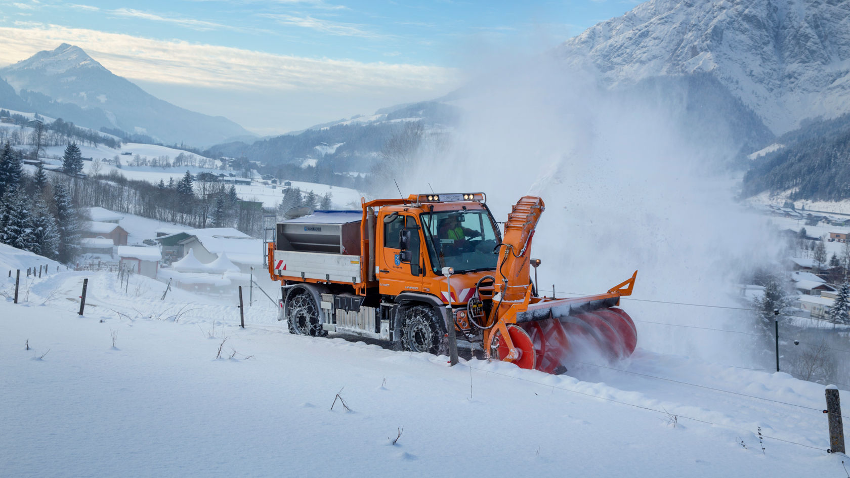 An orange Unimog with a snow blower clearing a snow-covered road. The snow blower throws a large amount of snow to the side, creating a cloud of snow. Another vehicle can be seen in the background in the snow.