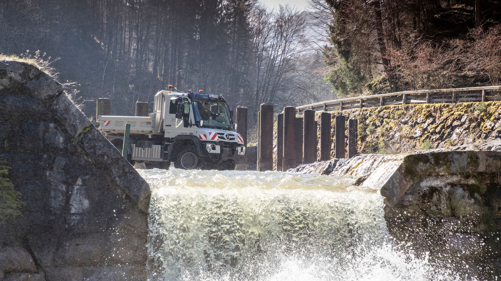 A Unimog crossing a small weir or waterfall. The water splashes around the wheels of the vehicle. The Unimog has a white cab and a grey loading surface. In the background is a wooded landscape. A stone wall and wooden posts frame the weir.