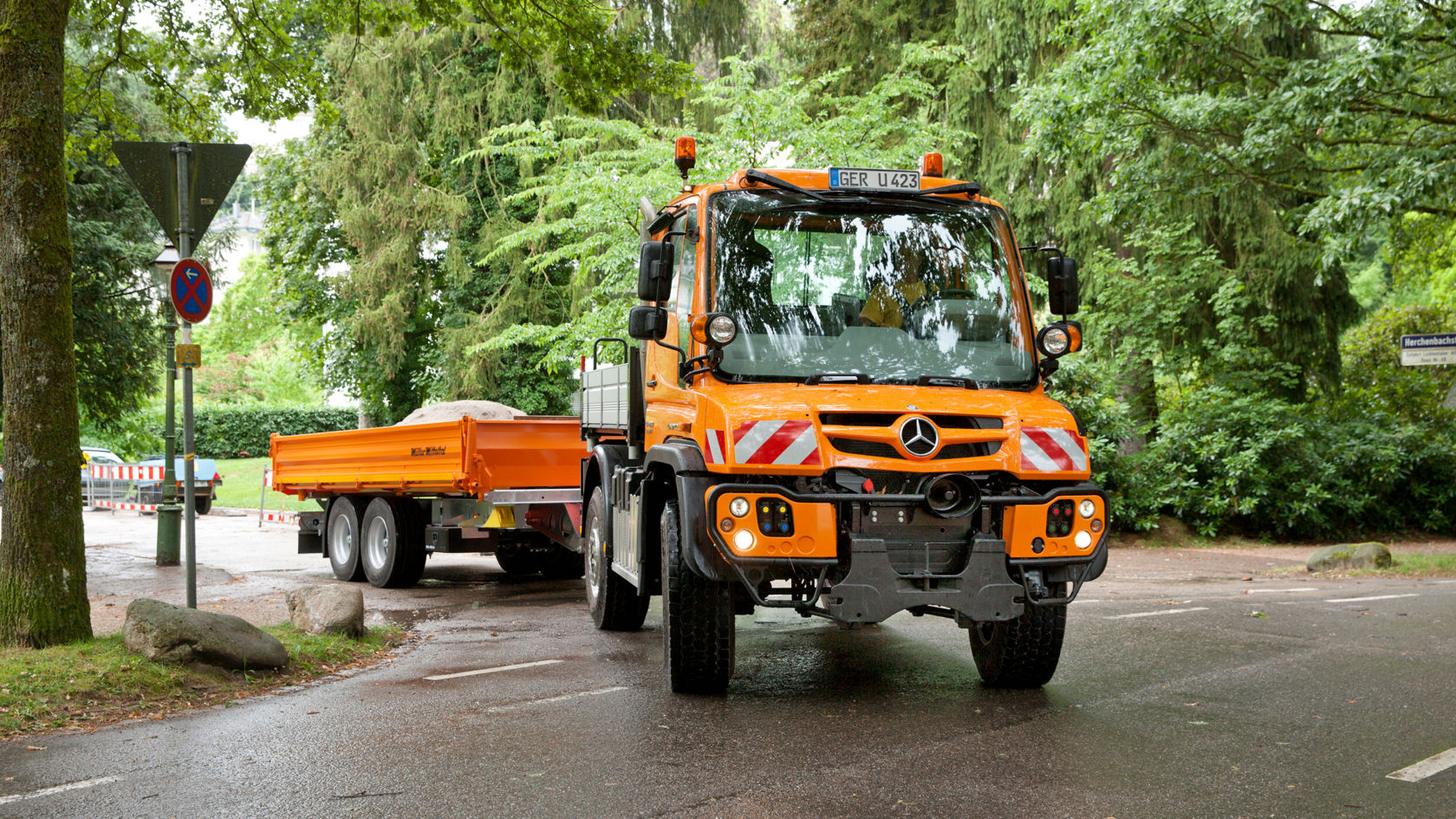 An orange Unimog pulling an orange flatbed trailer along an asphalt road. The truck is at a junction, surrounded by trees and shrubs. Other parked vehicles and traffic signs are visible in the background. The road surface seems to be damp.